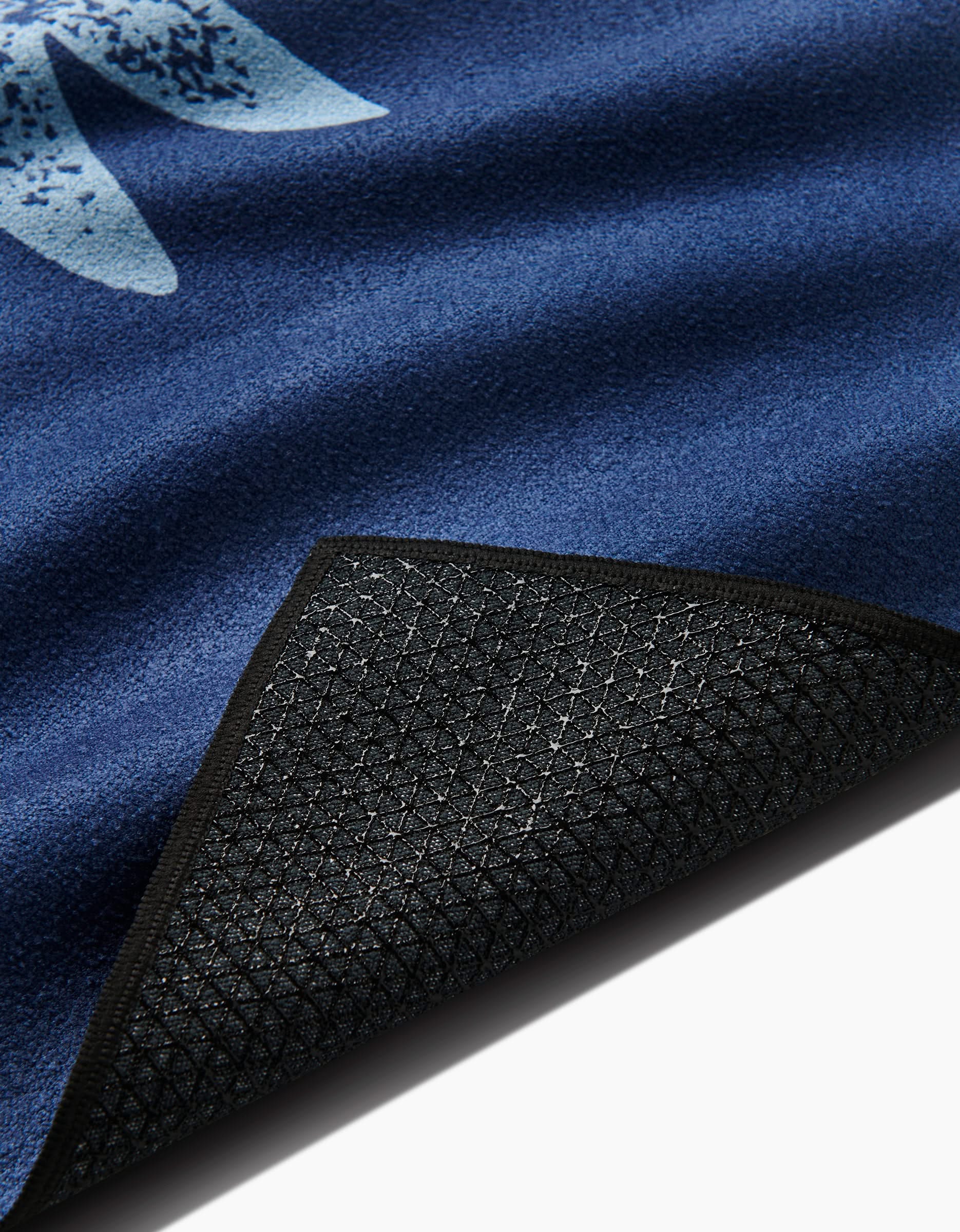 A close-up of the Blue Moon yoga towel by Graphics and Grain, featuring a blue textured surface with a light blue pattern and a flipped corner revealing its black, mesh-like slip-resistant backing for extra stability during practice.