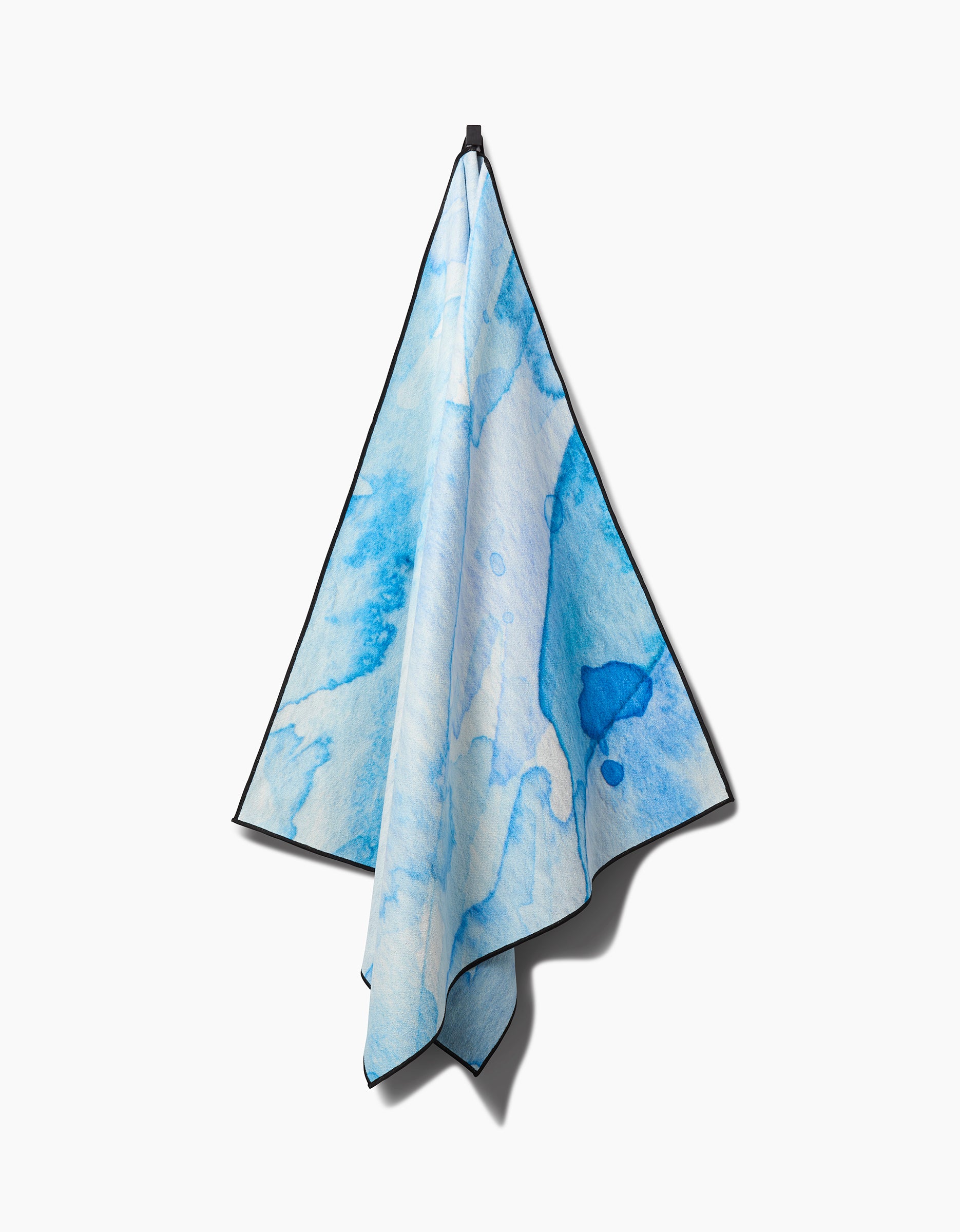 The GEOMETRY Boundless Blue yoga towel, showcasing an abstract blue watercolor pattern in various shades, hangs from a hook against a plain white background.