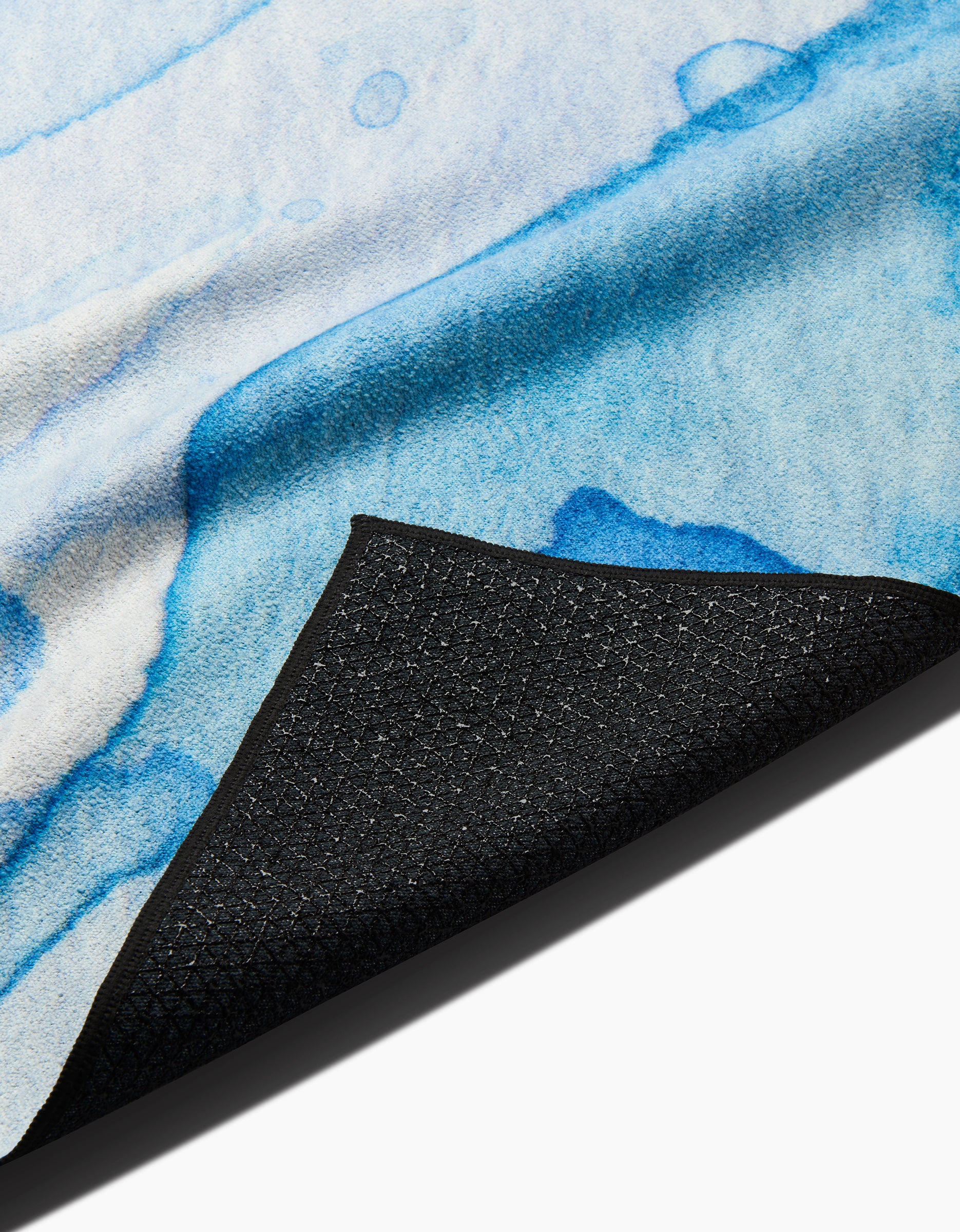 A close-up of the GEOMETRY Boundless Blue recycled yoga towel, featuring a blue and white watercolor design with a textured underside shown by flipping one black non-slip corner.