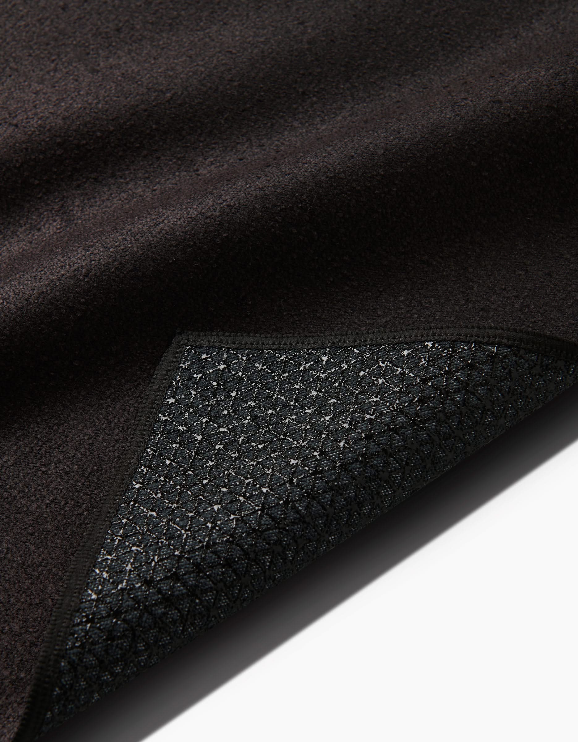 A close-up of the Cole Strem Brush Stroke yoga towel shows a dark, textured surface with a flipped corner, revealing its grid-patterned silicone grip underside. The top is soft, while the bottom has a slightly rough, rubberized feel.