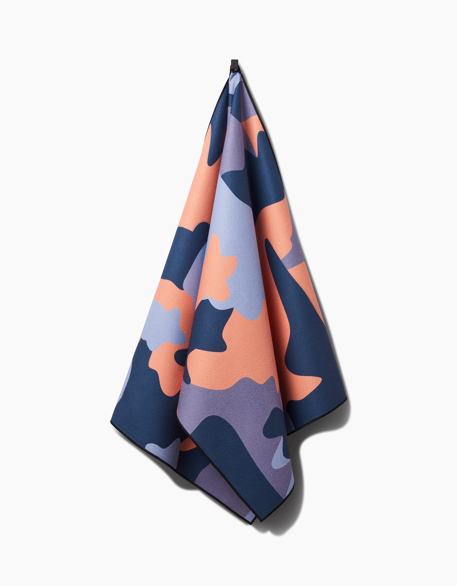 The GEOMETRY Camo yoga towel, featuring a slip-resistant design and a navy blue, peach, and lavender camouflage pattern, hangs against a white background.