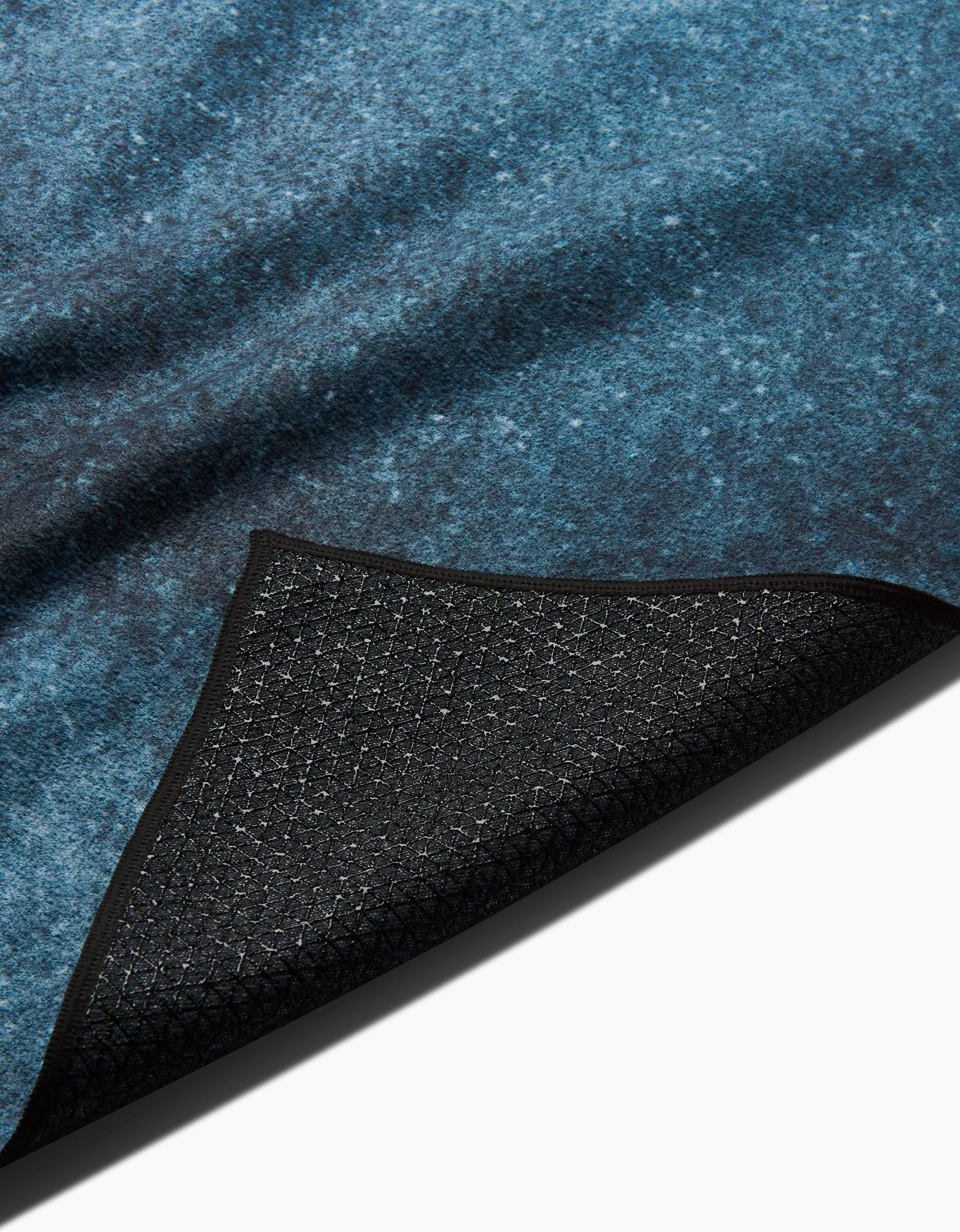 A close-up of the GEOMETRY Centered rug in blue, crafted from a recycled yoga towel. One corner is flipped to show its black diamond-patterned, non-slip backing. The rug is set against a white background.