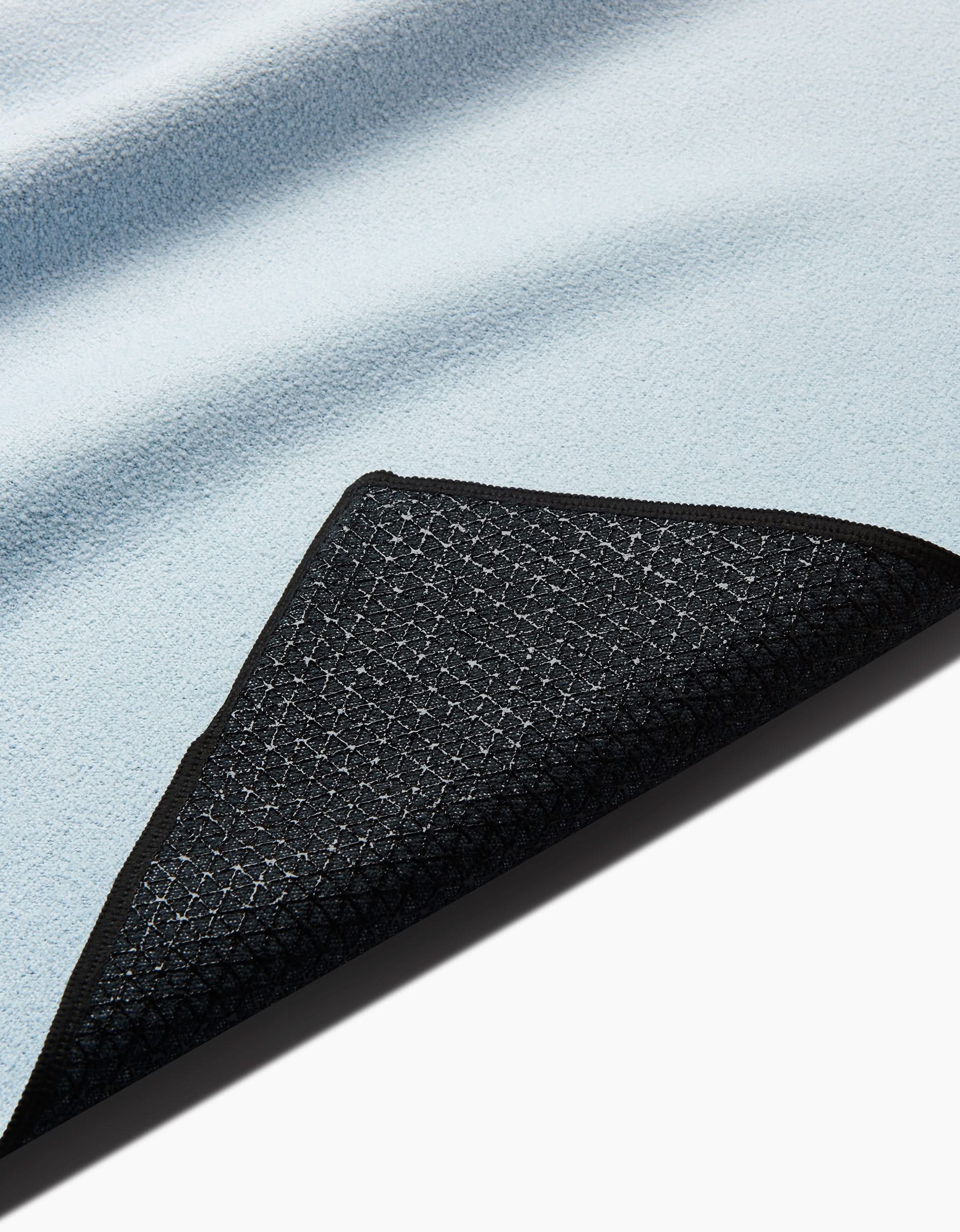 A close-up of the GEOMETRY Core yoga towel in light blue, with a folded corner showing its black, textured, grid-patterned underside and neatly finished edges. The towel is slip-resistant.