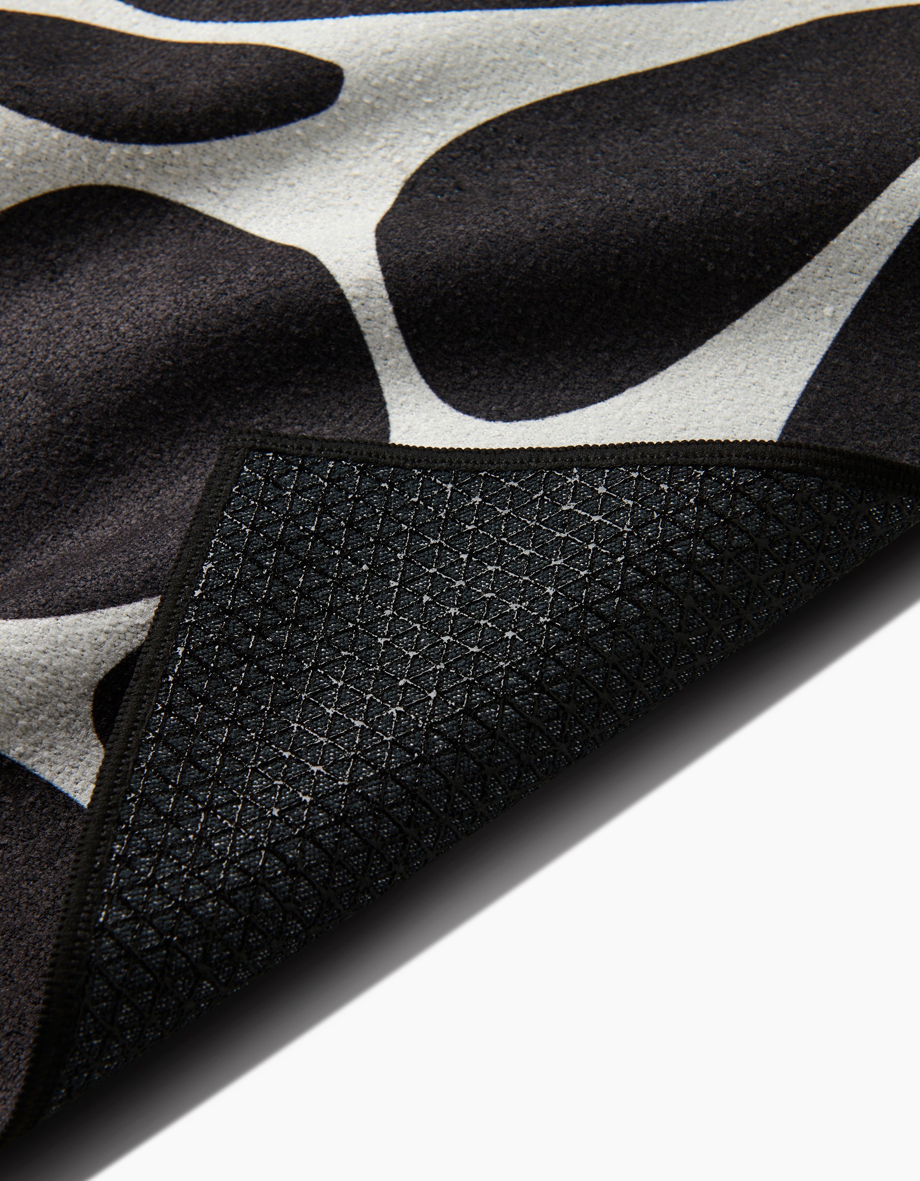 A close-up of the Cute Flowers rug by GEOMETRY shows its black and white pattern with a corner flipped to display the dark, textured, non-slip geometric grid backing, resembling a silicone grip yoga towel.