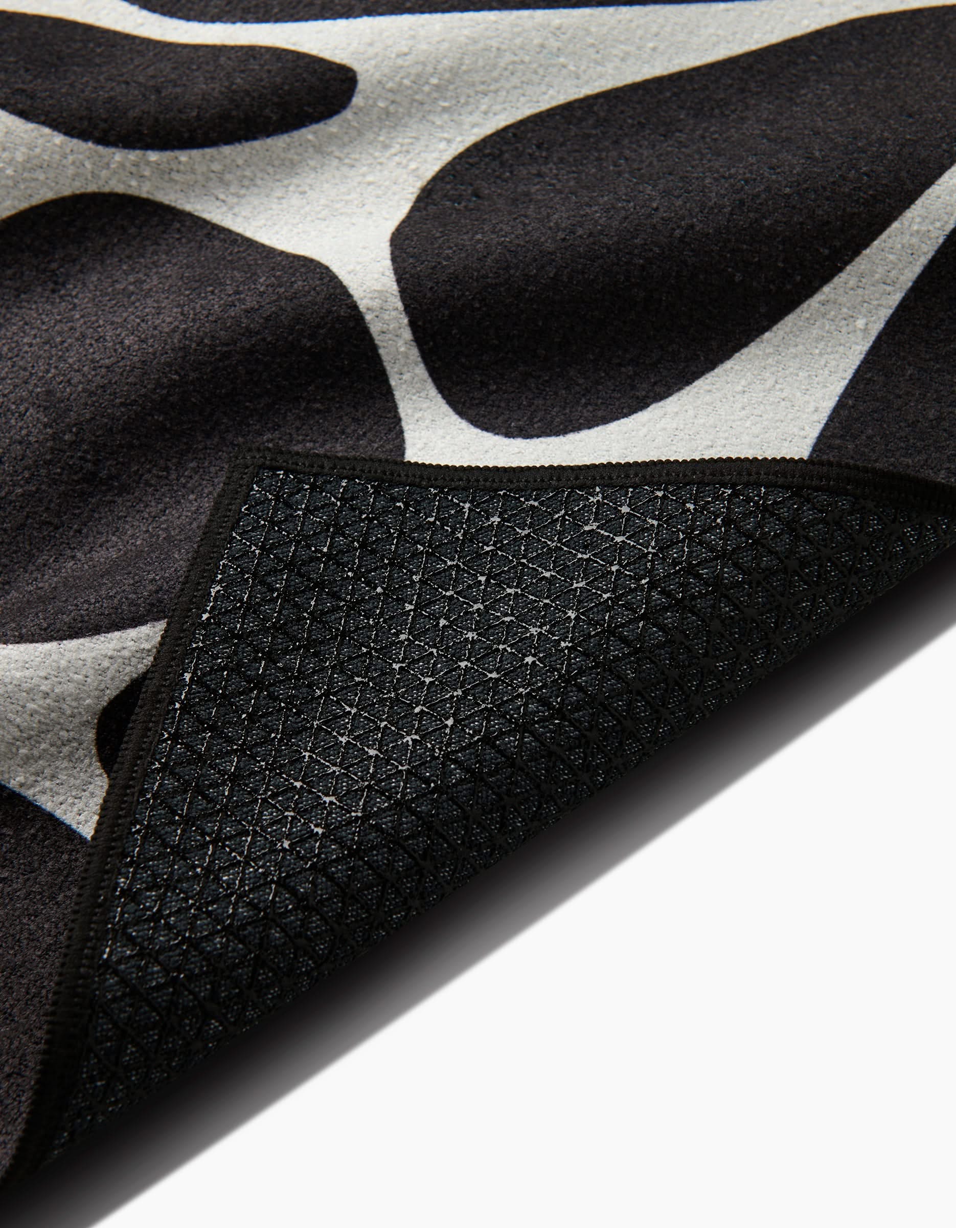 A close-up of the Cute Flowers rug by GEOMETRY shows its black and white pattern with a corner flipped to display the dark, textured, non-slip geometric grid backing, resembling a silicone grip yoga towel.