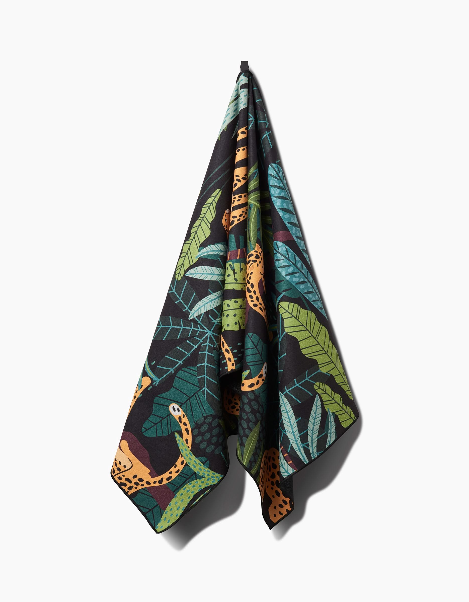 The GEOMETRY Feeling Spotty yoga towel, made from recycled materials, features green tropical leaves and orange cheetahs on a black background and hangs stylishly on a hook.
