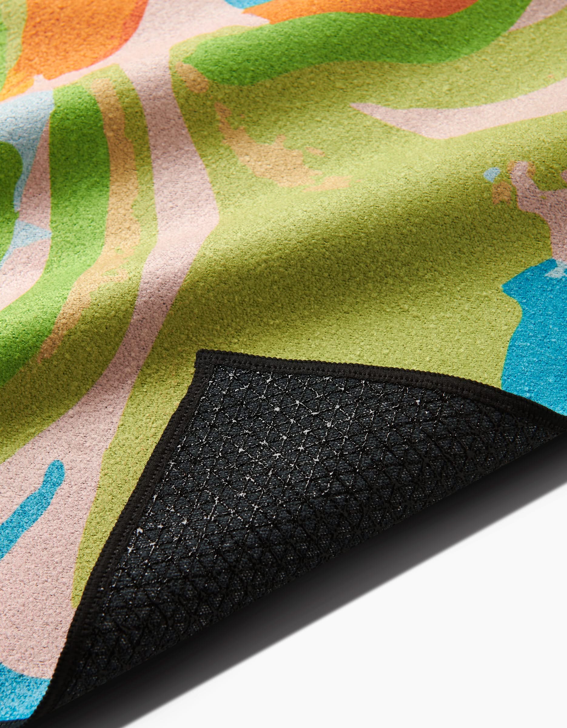 A close-up of Sareka Unique’s Florescence rug showcases abstract green, orange, pink, and blue patterns, with a flipped corner revealing a textured black non-slip backing made from recycled materials.