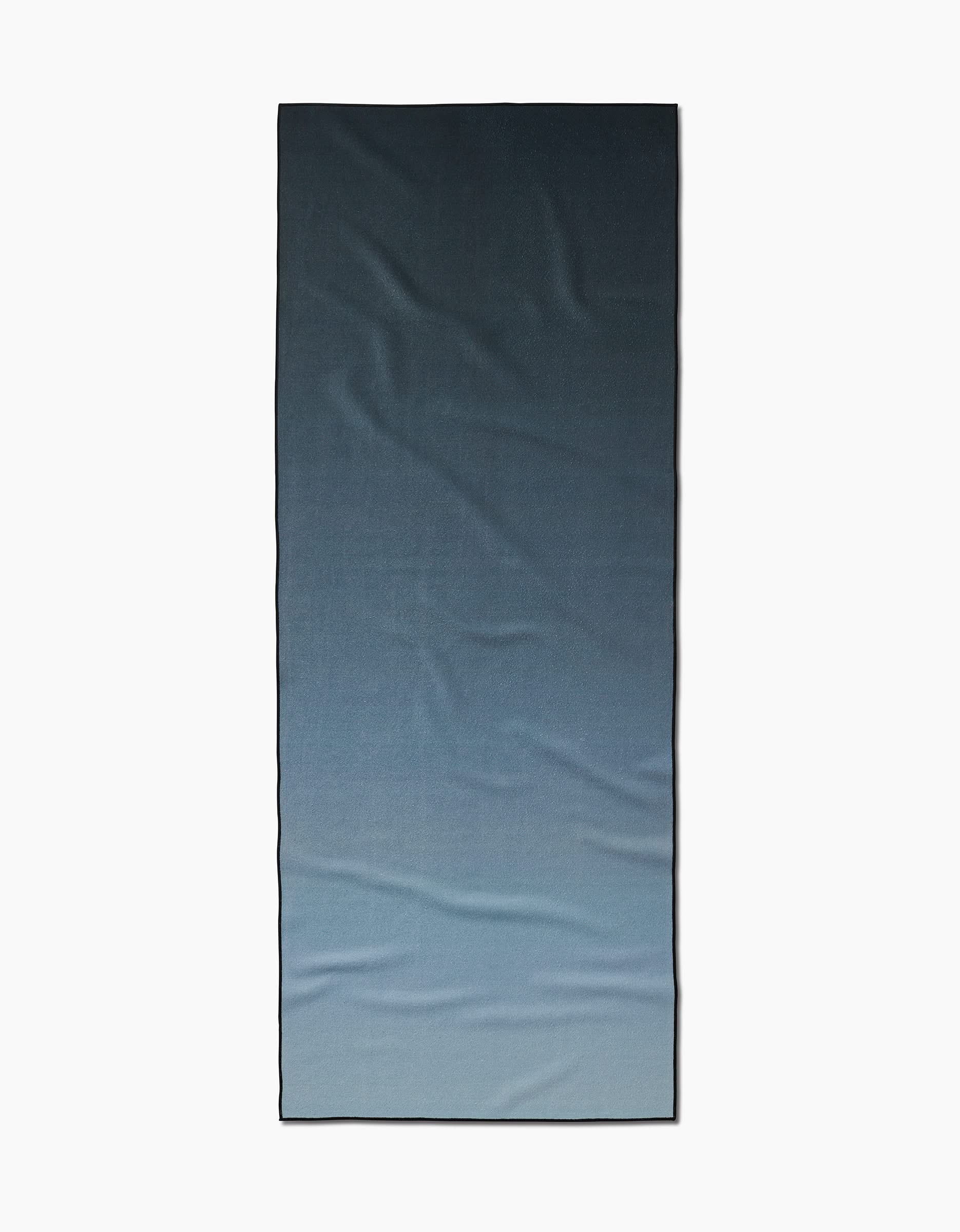 The Foundation yoga towel is rectangular, made from recycled materials, and displays a smooth gradient from dark blue at the top to light blue at the bottom. It is pictured flat on a white background.