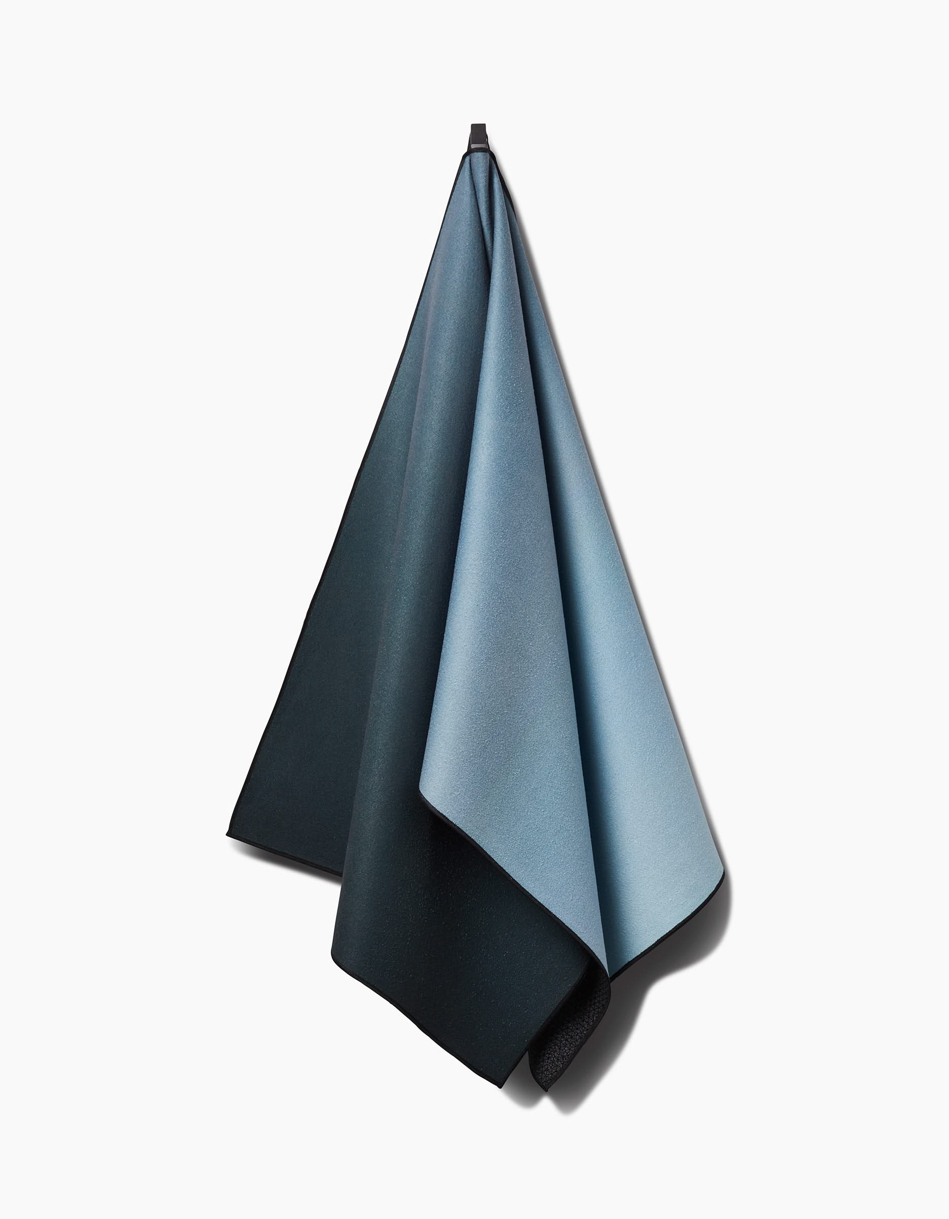 The Foundation yoga towel, made from recycled microfiber, hangs from a hook against a white background. Its color transitions seamlessly from light blue to dark gray and the fabric drapes naturally.