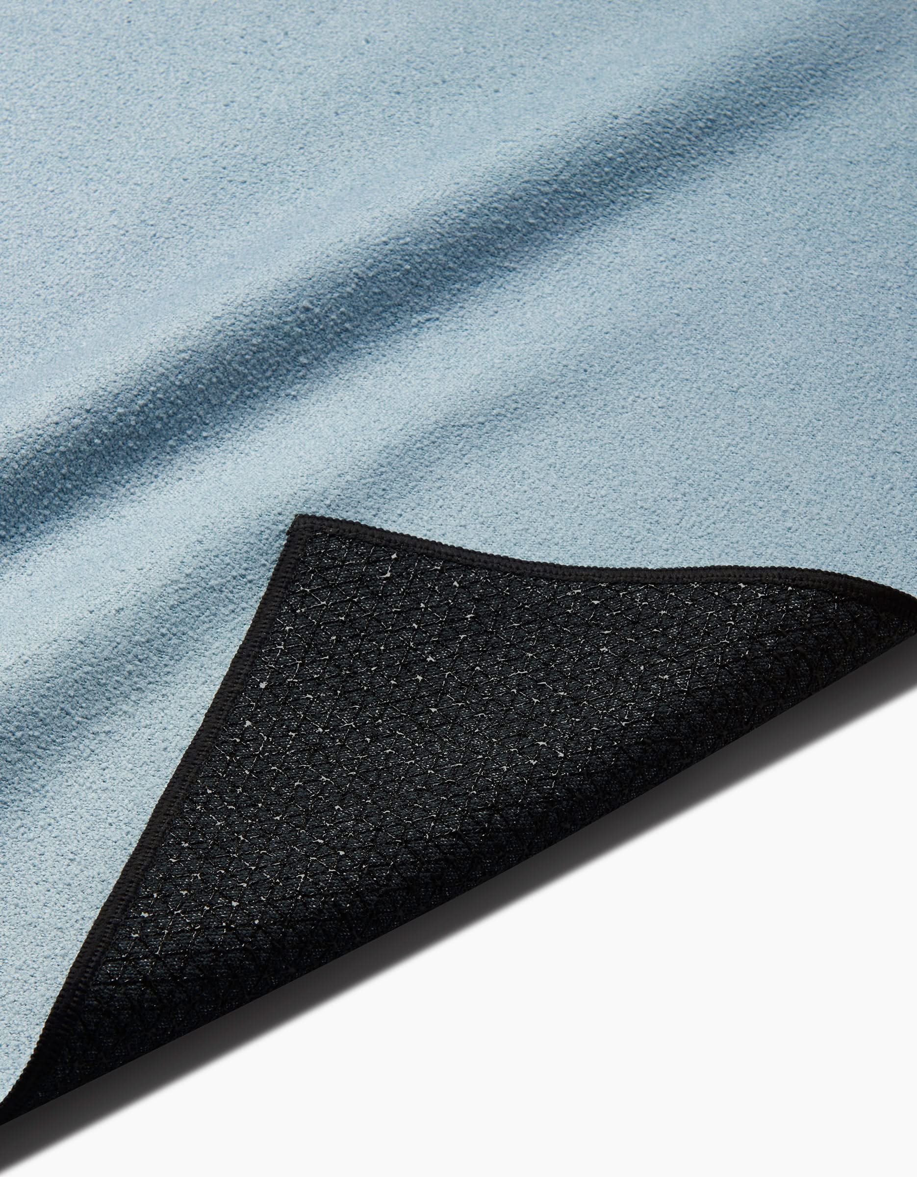 A close-up of the Foundation yoga towel in light blue, featuring textured fabric and a black slip-resistant backing, with one corner folded to reveal the underside.