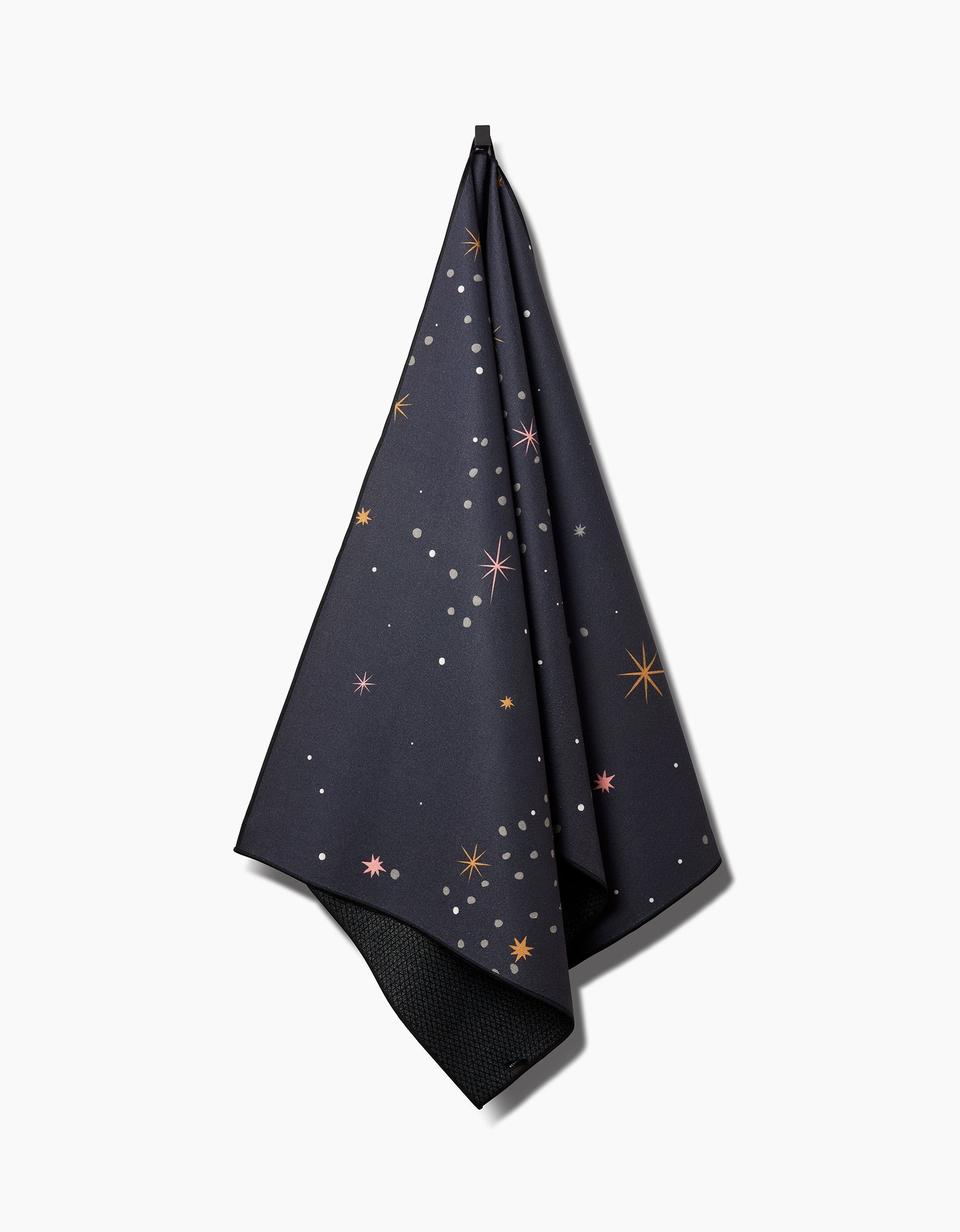 The Galaxy Burst yoga towel by Ash Cascade hangs from a hook, showcasing its dark fabric adorned with pastel stars and dots that evoke a night sky.