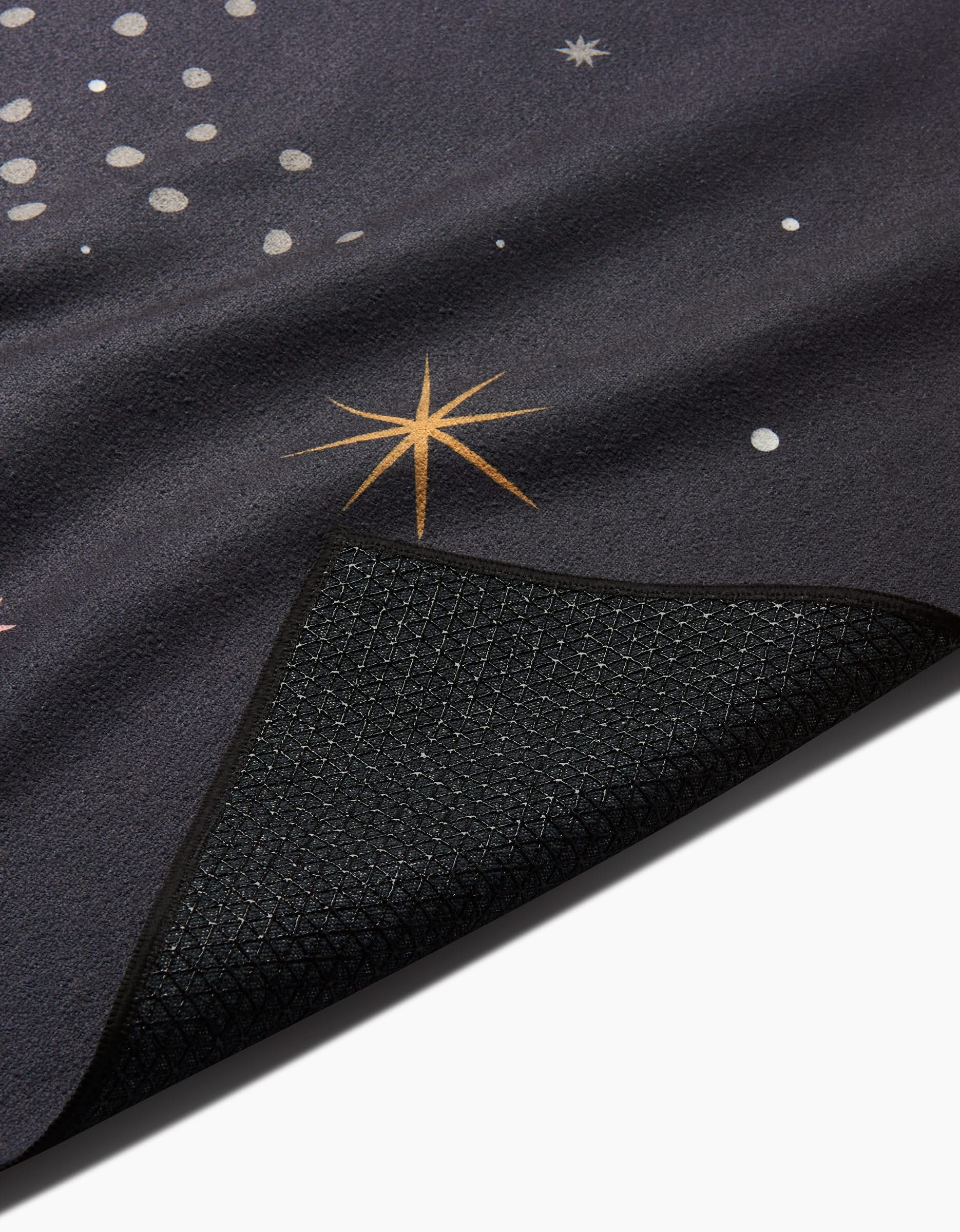 A close-up of Ash Cascade’s Galaxy Burst yoga mat features star and dot patterns, with a corner flipped to reveal its textured black, nonslip underside—ideal for use with your favorite non-slip yoga towel.