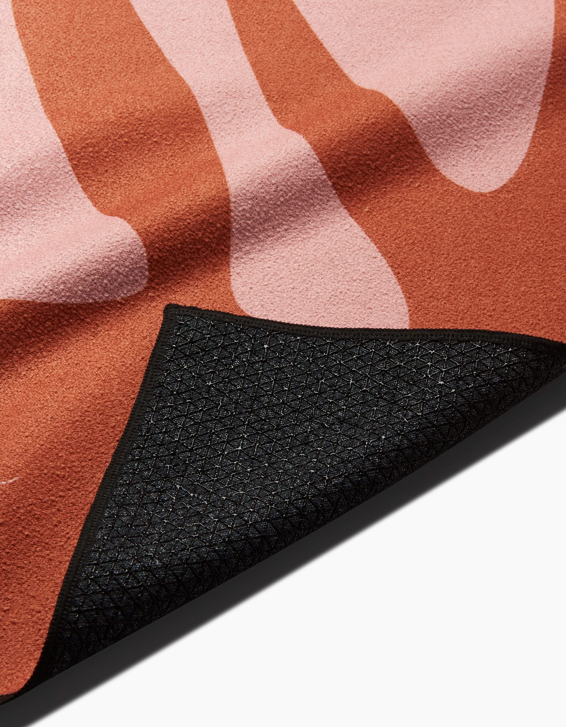 A close-up of the GLORIOUS mat by Arielle Estoria shows pink and rust abstract shapes. One corner is flipped, revealing a black non-slip yoga towel backing with a subtle geometric pattern.