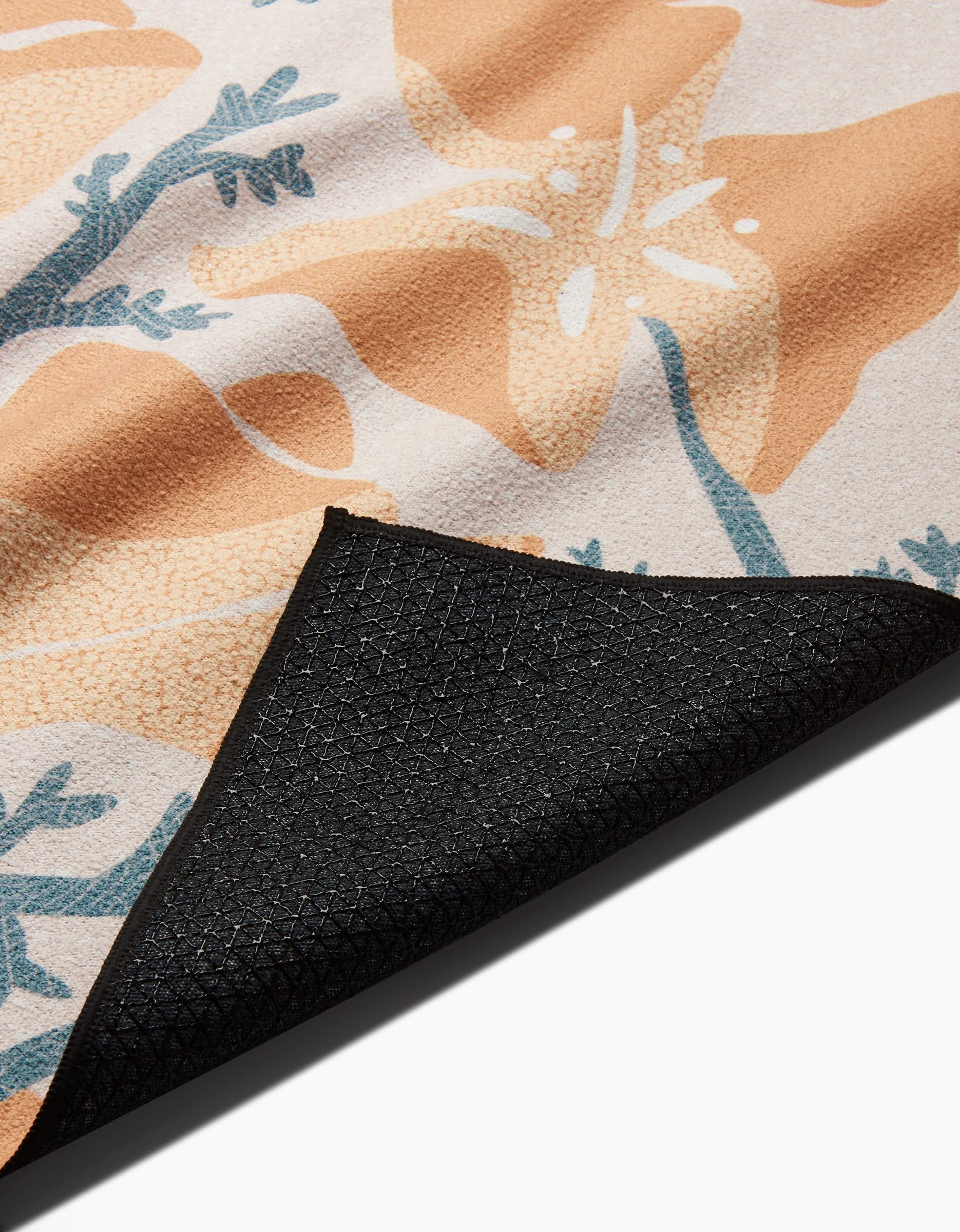 A close-up of Julianne Haness’s Golden Poppy recycled yoga towel shows its peach, teal, and beige floral pattern with one corner folded to reveal the textured, slip-resistant black underside.