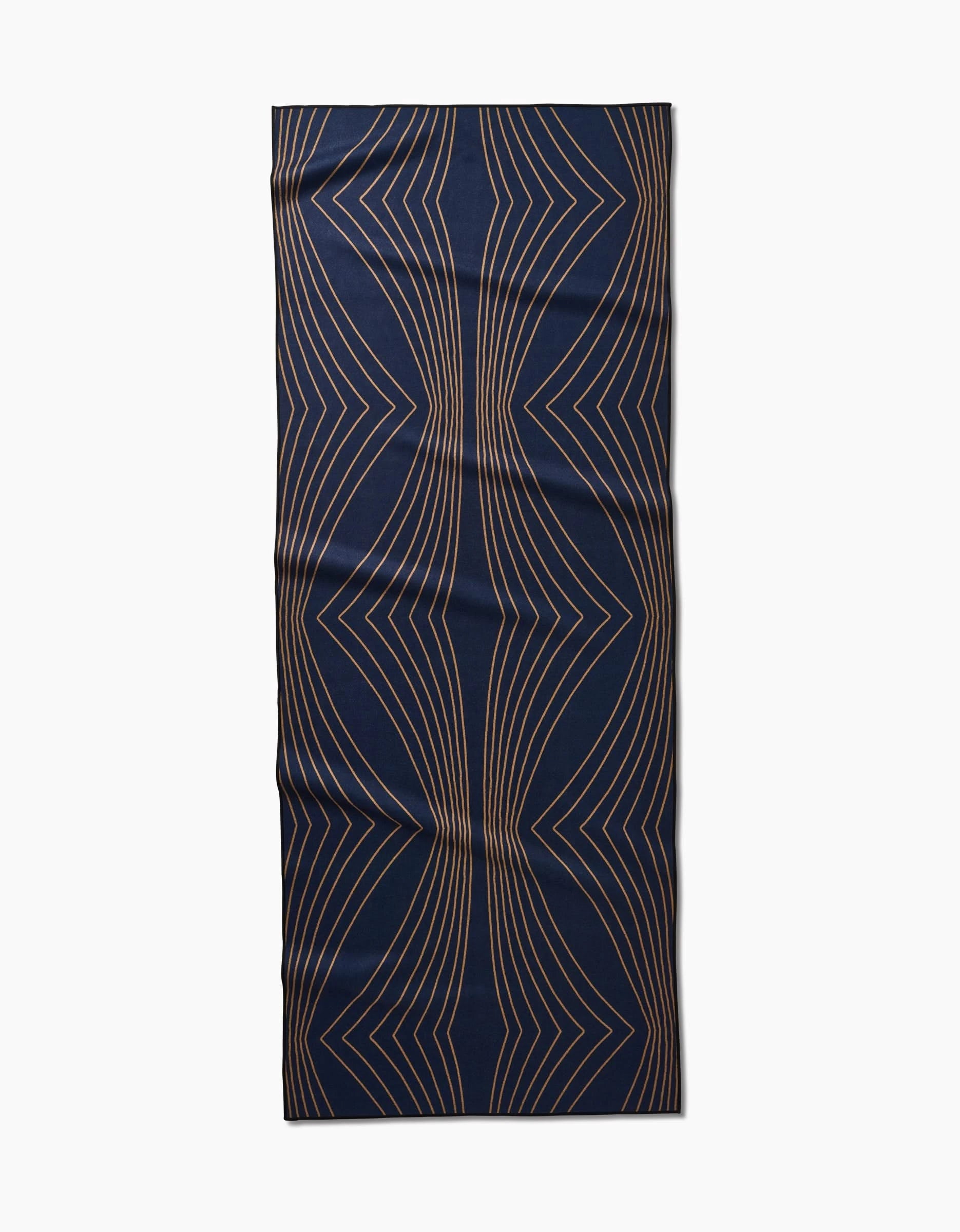 The Hera yoga towel by Amy MacCready features a rectangular, dark blue non-slip surface with curved, symmetrical gold geometric lines in a wavy pattern. It is shown lying flat on a white background.