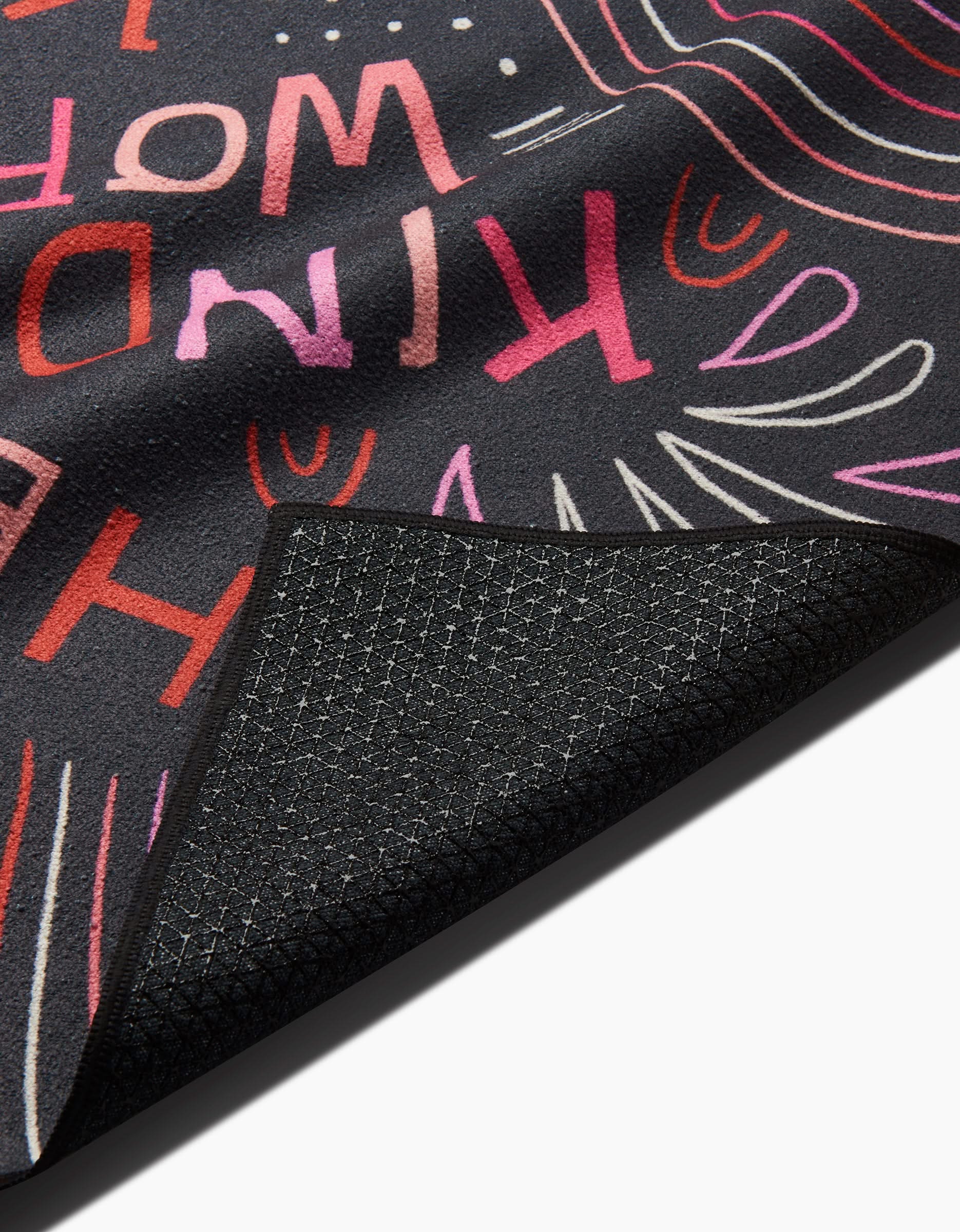 A close-up of the Jessica Swift Kind Hearts yoga mat shows colorful abstract lines and the partially visible phrase KIND WORLD in bold pink and red, with a corner flipped to reveal its textured underside—ideal with a slip-resistant towel.