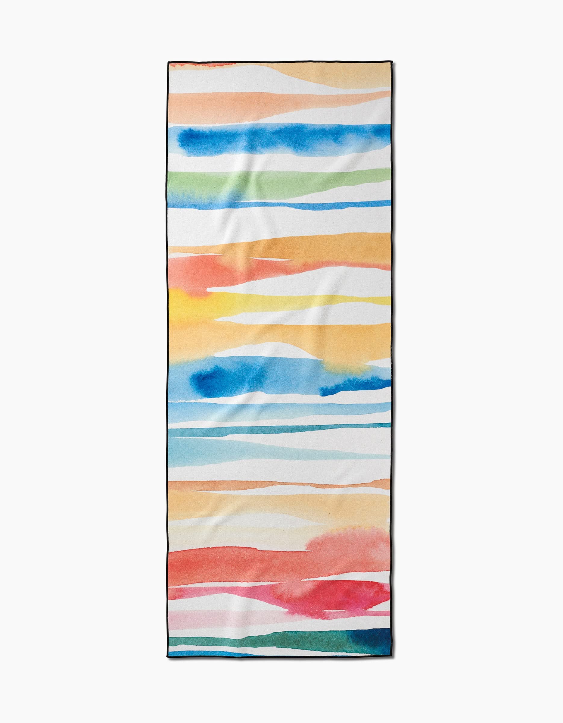 The Sara Mari Lakeside yoga towel is rectangular, made from recycled materials, and features horizontal watercolor stripes in blue, orange, green, yellow, and red on a white background.