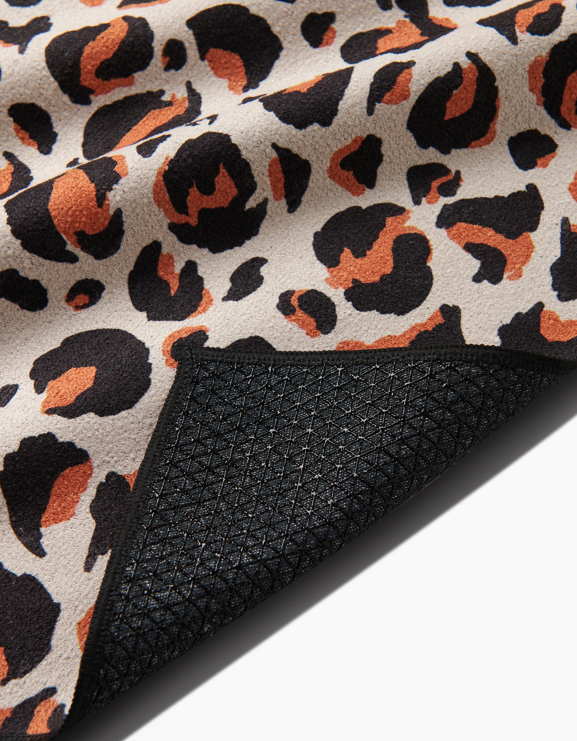 A close-up of the GEOMETRY Leopard yoga towel, made from recycled materials with a black, orange, and beige leopard print. A corner folds back to show the black silicone grip textured backing.