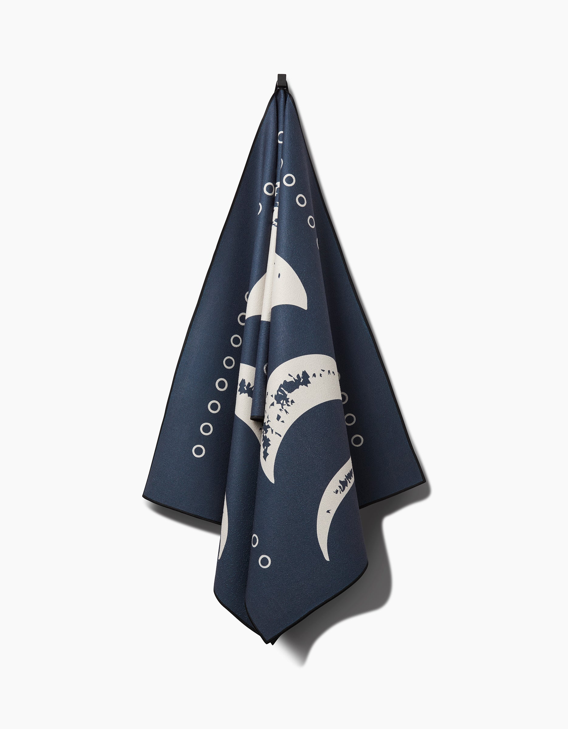 The Lunar Phases yoga towel by Ashley Celena, made from recycled materials, features a navy blue background with white crescent moons and small circles, and hangs from a hook set against a plain white background.