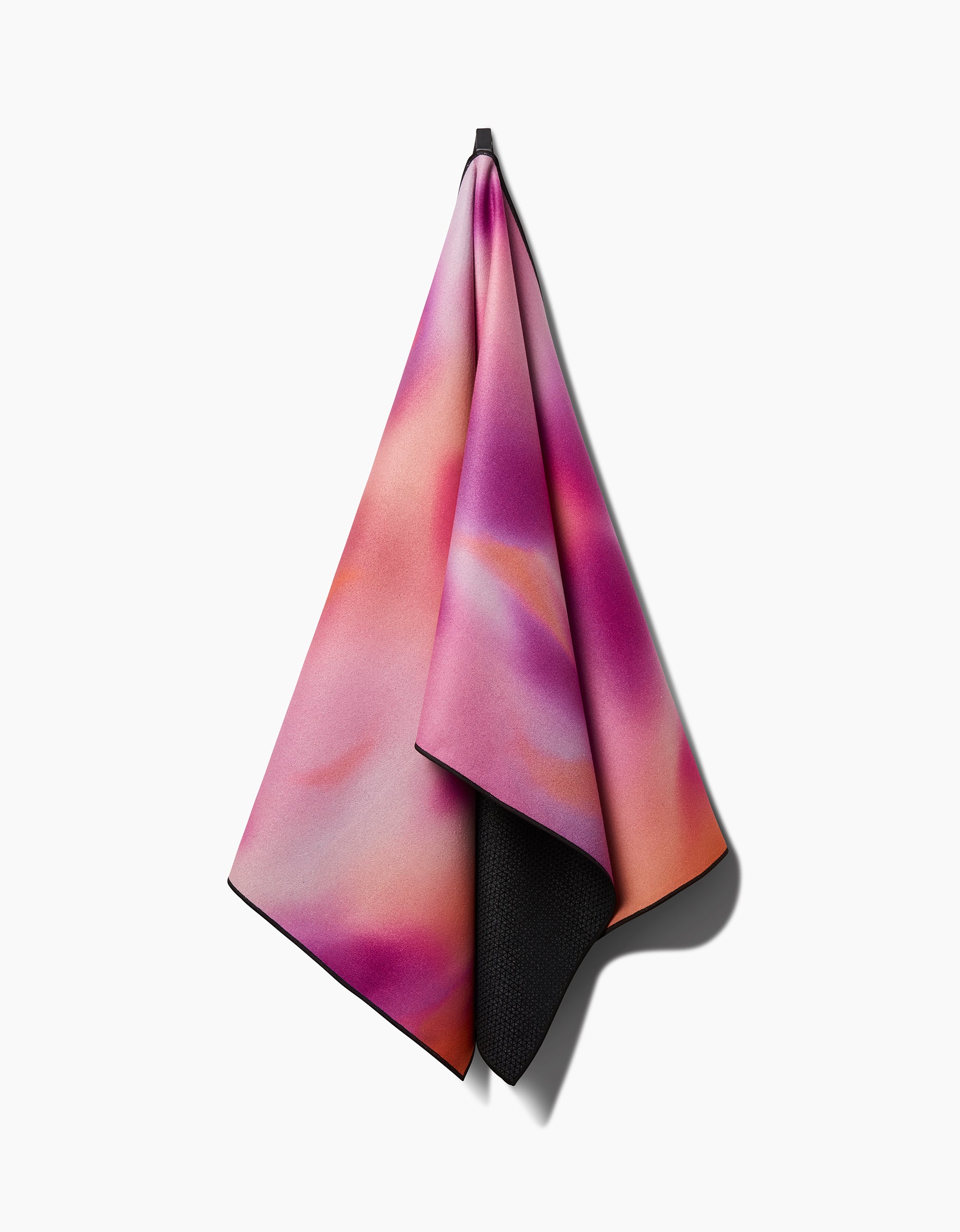 The GEOMETRY Magenta Yoga towel features a soft, abstract pink, purple, and orange pattern. Hanging from a hook against a white background, its black textured silicone grip underside is visible at the edge.
