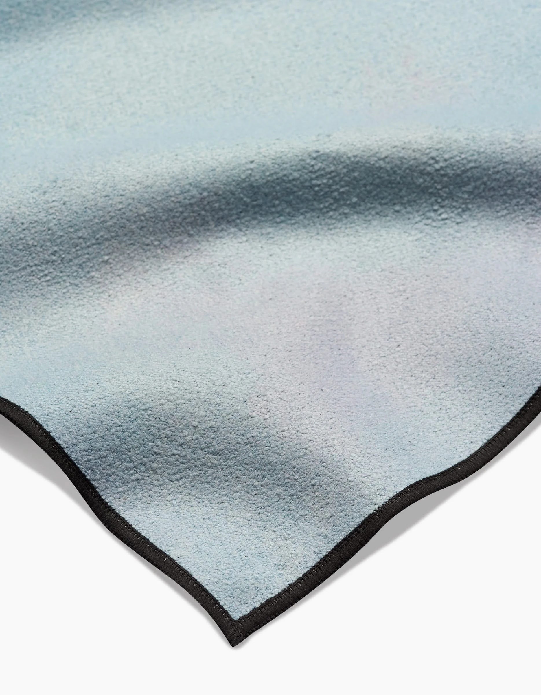 A detailed view of Stefanie Bales’ Magic in the Sky yoga towel, crafted from recycled materials in a soft light blue hue, showcasing a black smooth-edged border, gentle folds, and a subtle color gradient.