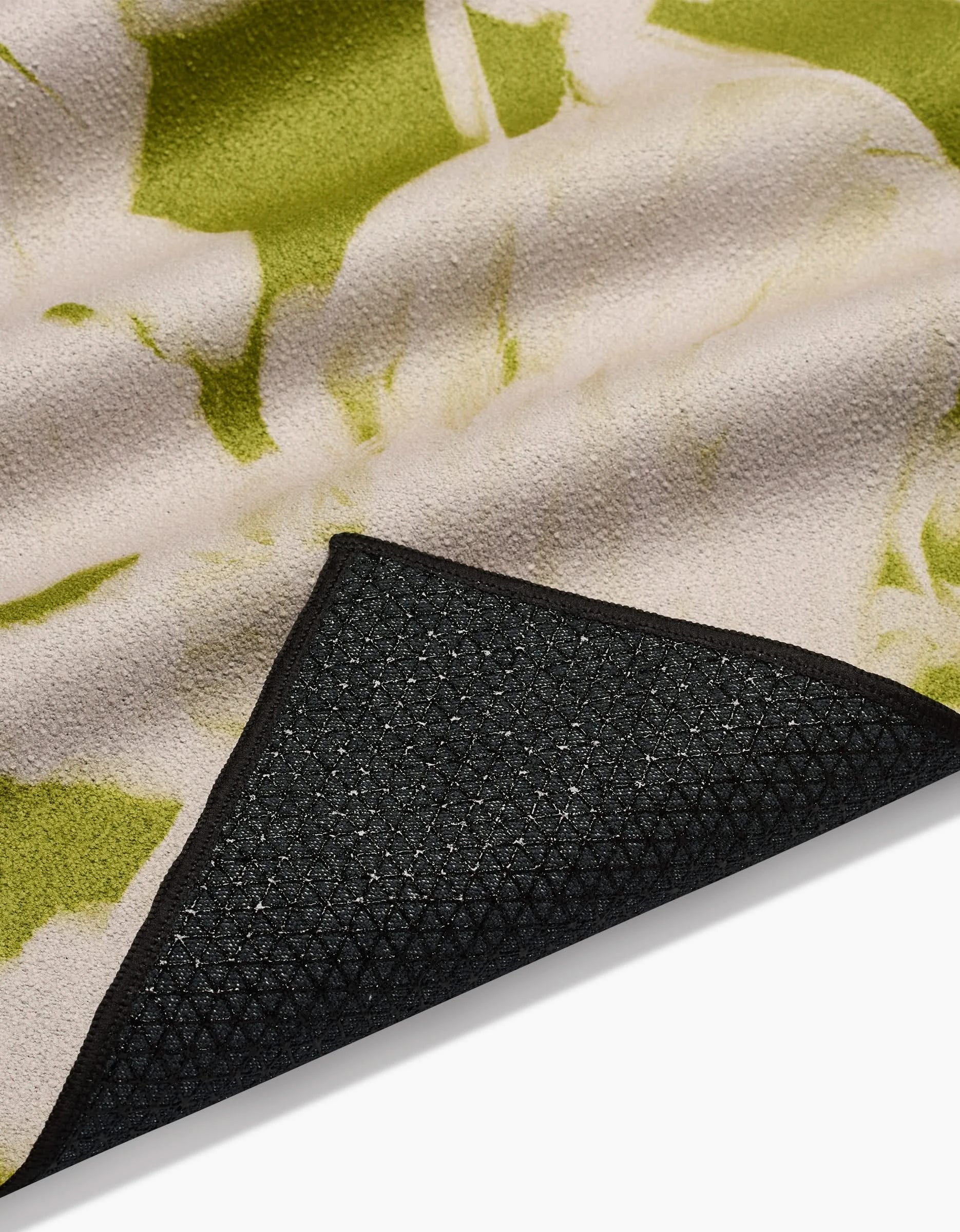 A close-up of GEOMETRYs Meadow Mist yoga towel shows its green and beige abstract pattern and black textured silicone grip non-slip backing, partially folded to reveal the underside—crafted from recycled materials.