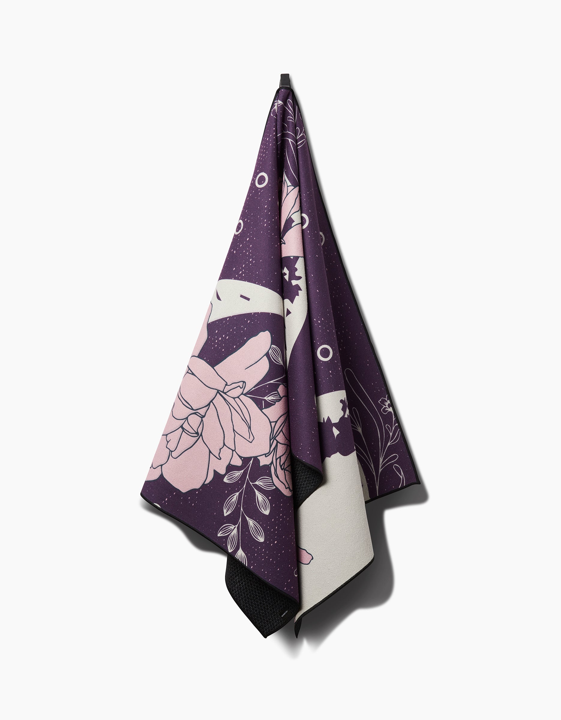 The Ashley Celena Midnight Garden yoga towel, featuring slip-resistant fabric with pale pink floral and abstract designs, hangs on a hook against a white background.