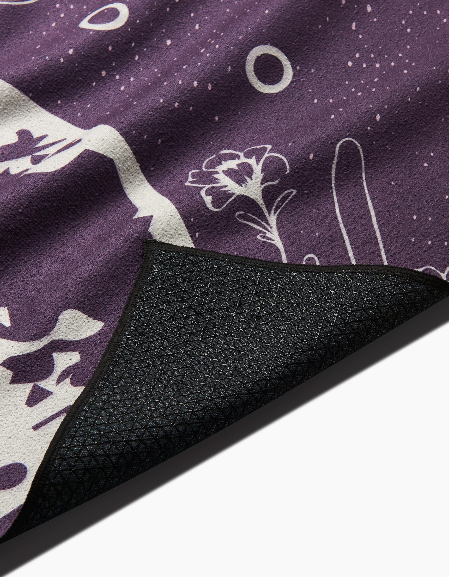 A close-up of the Ashley Celena Midnight Garden yoga towel, showcasing purple fabric with white floral and abstract patterns and a curled textured black slip-resistant backing.