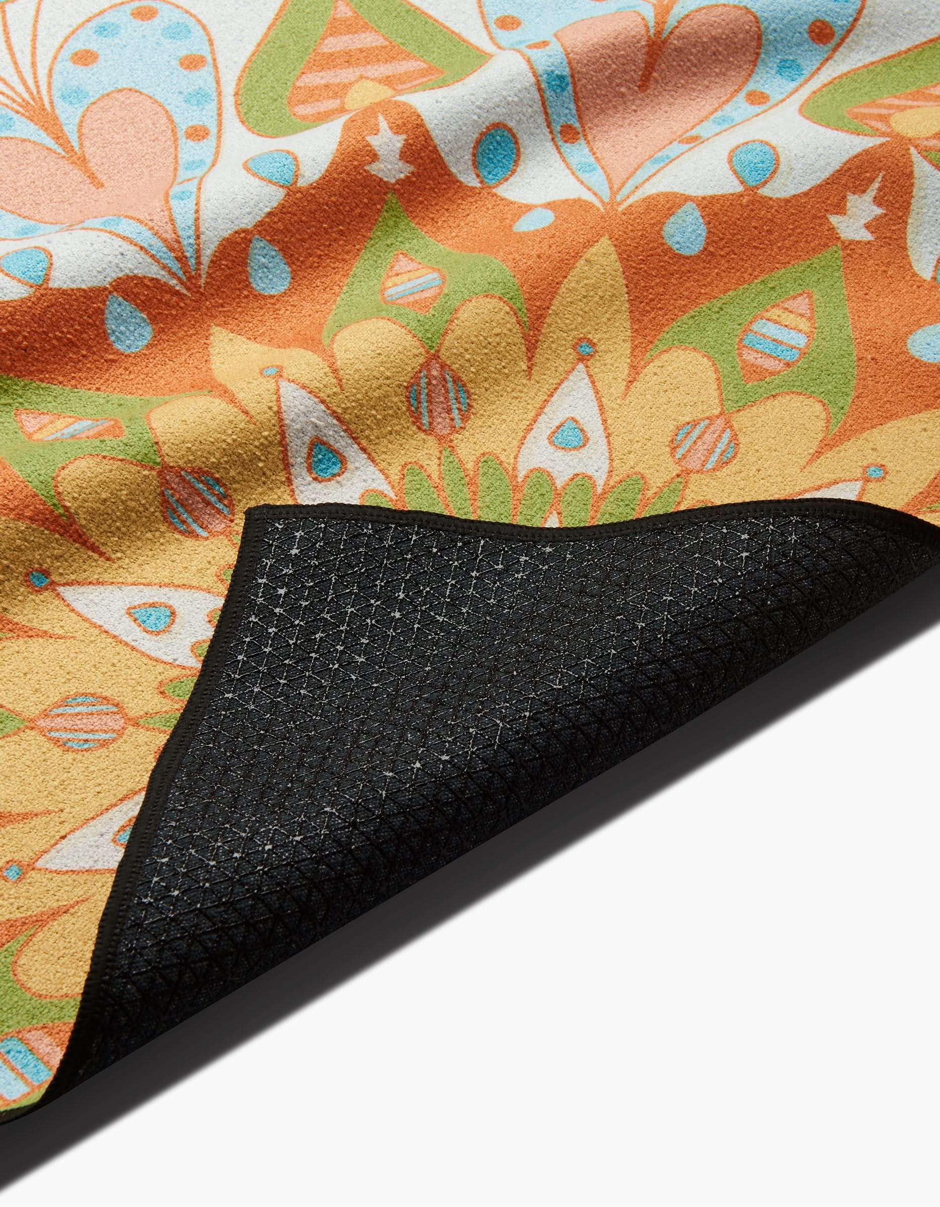 A close-up of the Mindfulnice Mindful Mandalas yoga towel, featuring vibrant orange, yellow, green, and blue mandala designs, with its textured black non-slip silicone underside folded over for grip visibility.