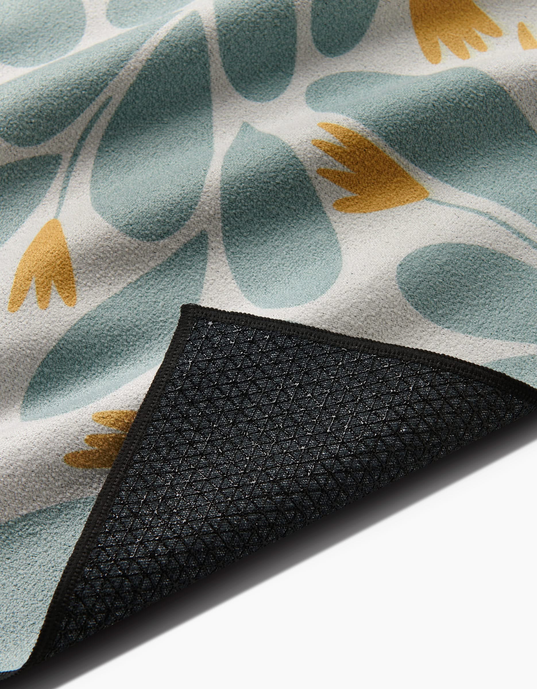 A close-up of the Mustard Pinwheel rug by Rebecca Woolbright, made from recycled materials with blue leaves and yellow flowers on a light background; one corner reveals its black, textured non-slip backing.
