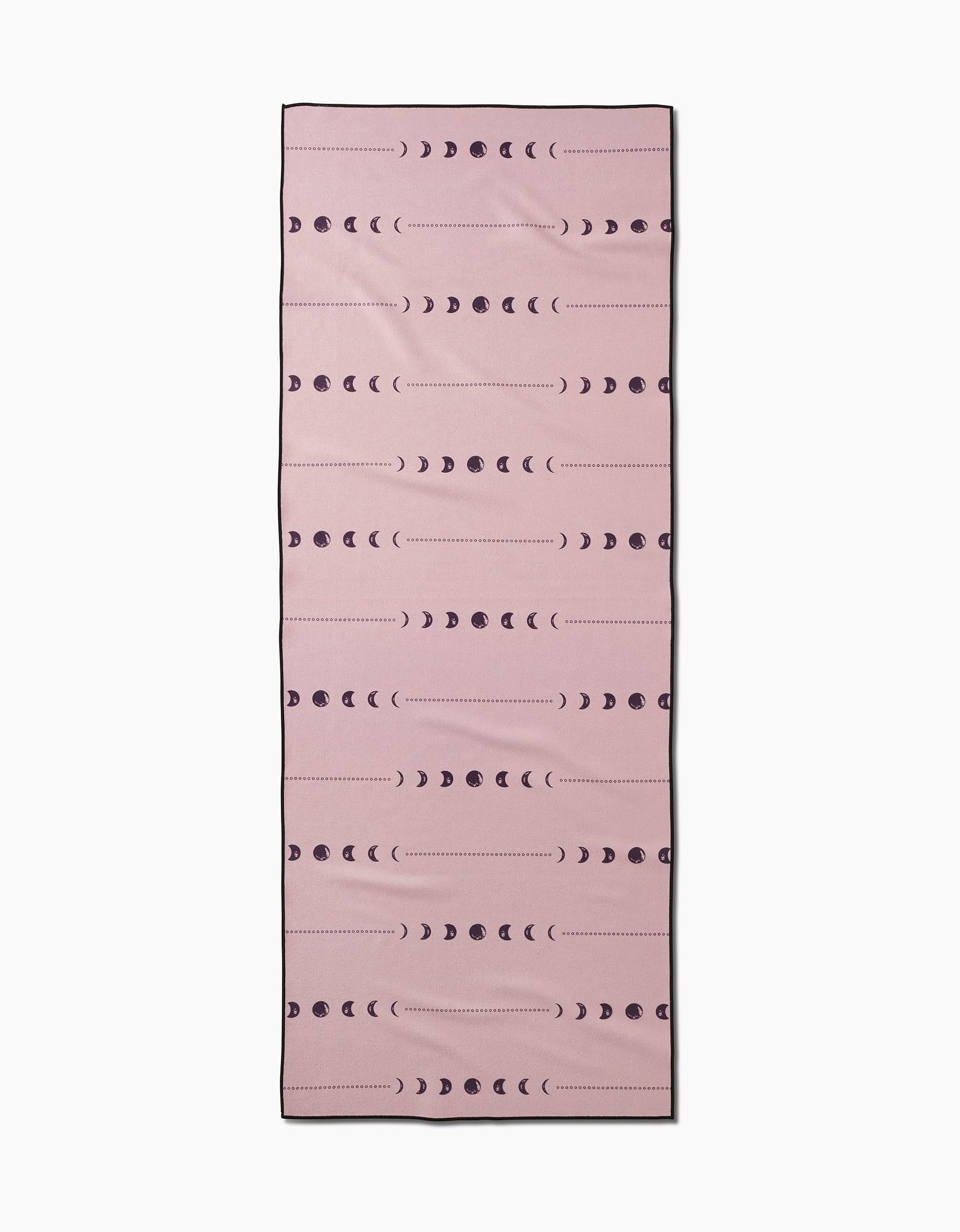 The Night Sky yoga towel by Ashley Celena is rectangular and pink, featuring rows of dark moon phases in crescents and circles. Silicone grip backing provides extra stability during your practice.