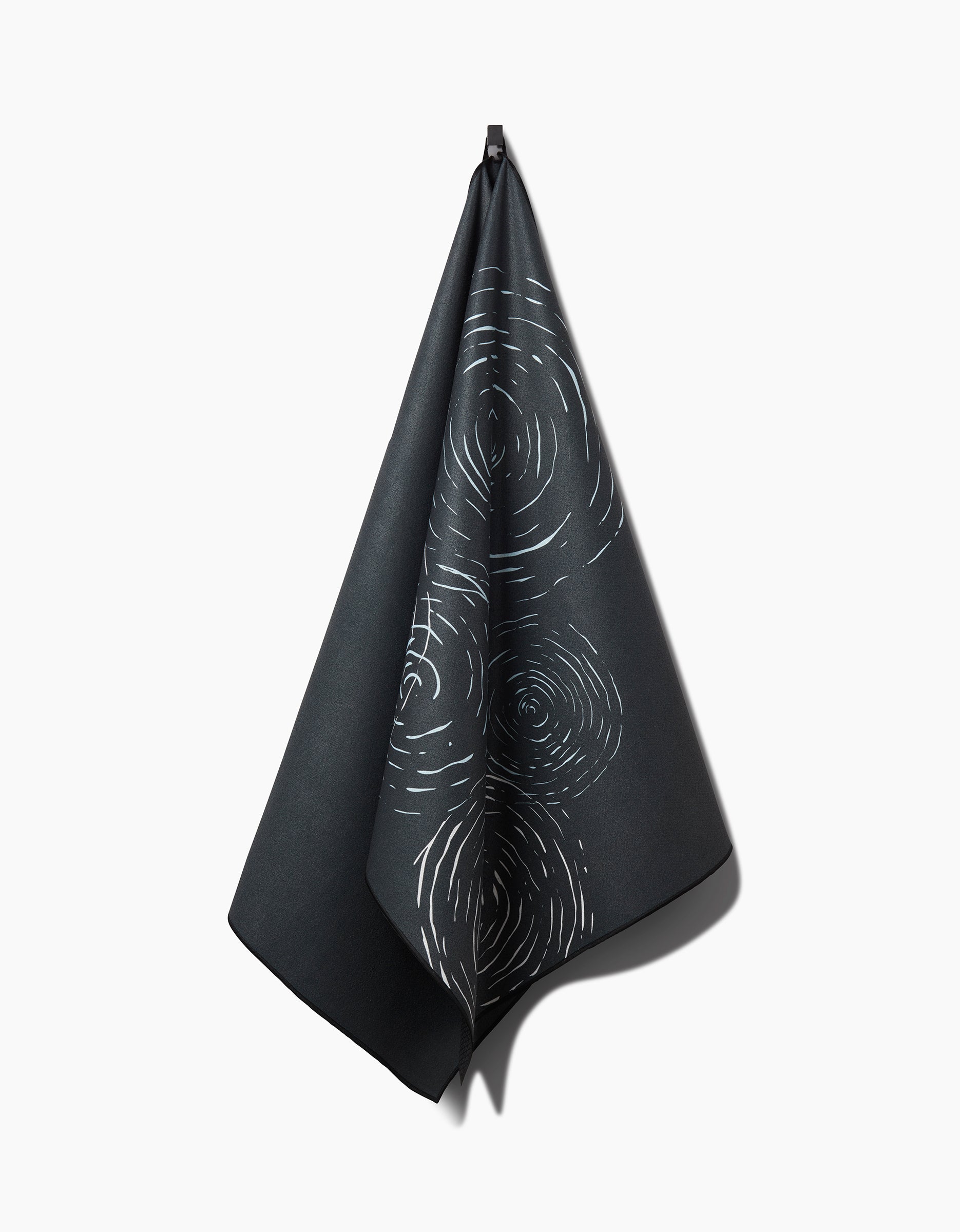 The GEOMETRY Om yoga towel, in a dark hue with concentric white lines like tree rings or ripples, hangs from a hook against a plain white background.