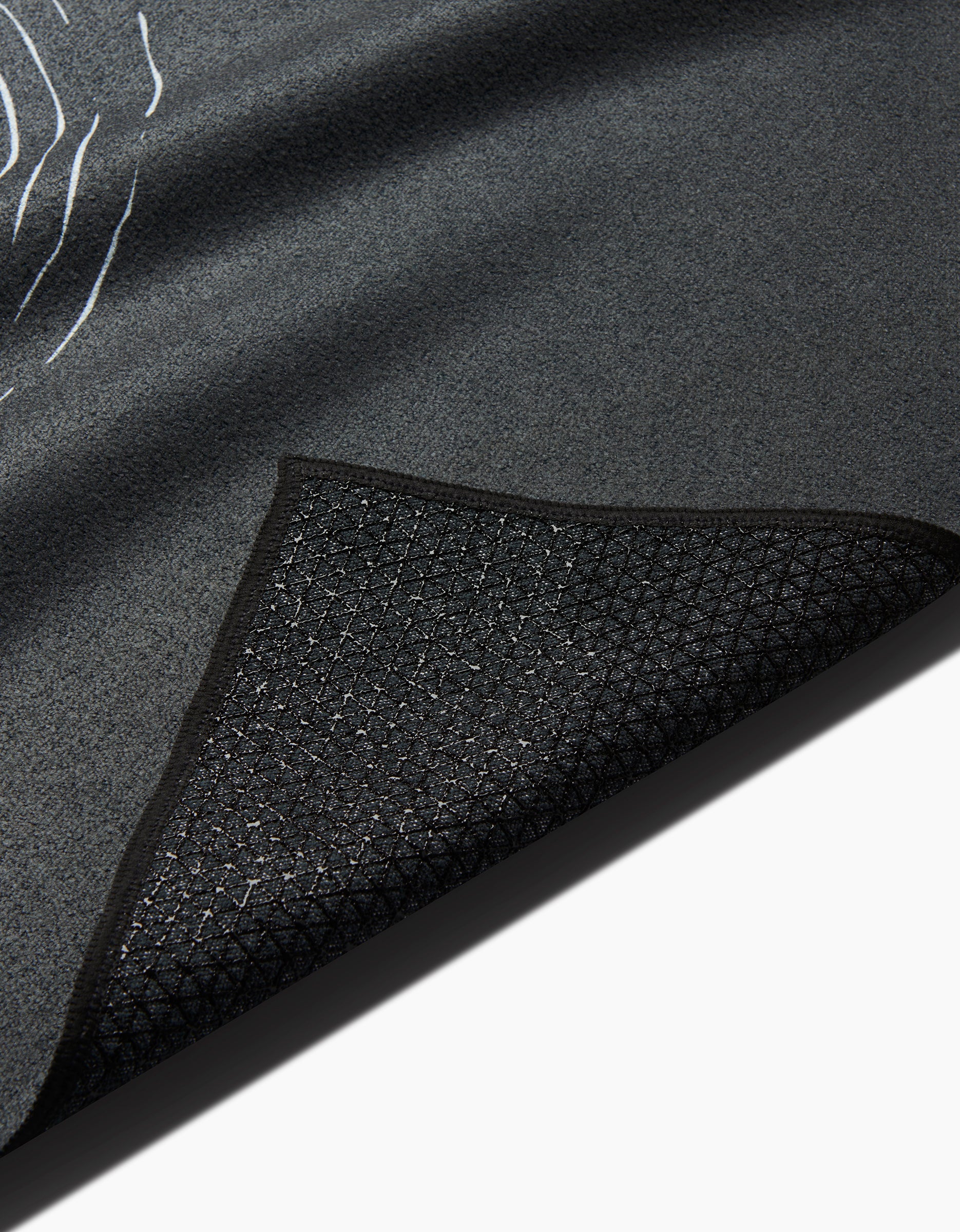 A close-up of the GEOMETRY Om black yoga towel shows its textured surface with one corner flipped, revealing a diamond-patterned silicone grip underside against a white background.