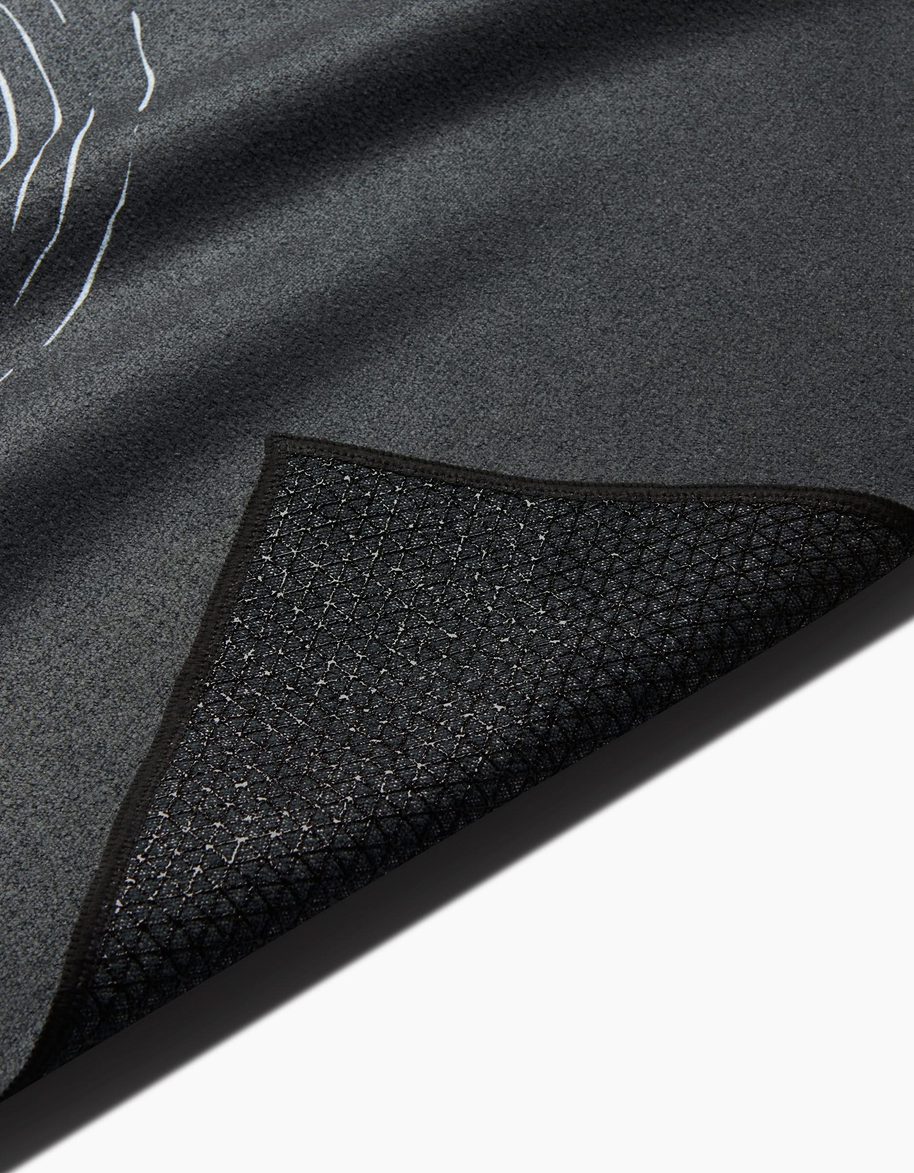 A close-up of the GEOMETRY Om black yoga towel shows its textured surface with one corner flipped, revealing a diamond-patterned silicone grip underside against a white background.