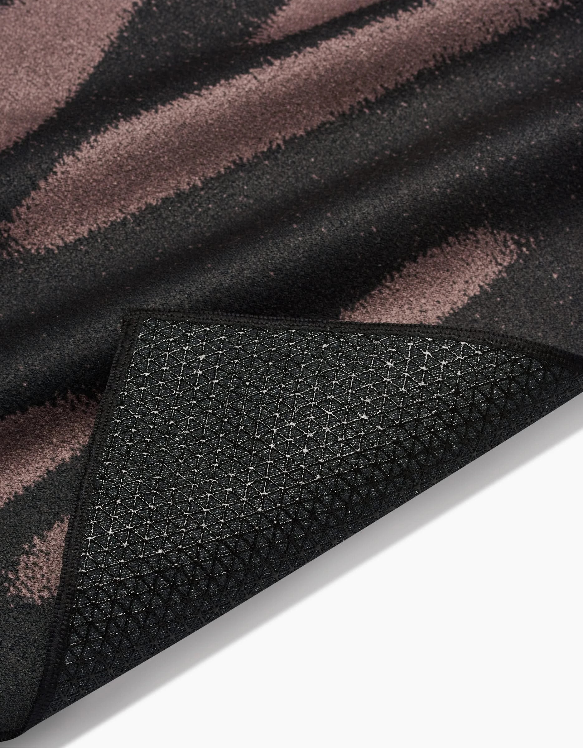 A close-up of GEOMETRYs Palm Shadow rug corner, flipped to reveal its black textured slip-resistant backing. The surface displays black and muted mauve brush strokes, photographed against a white background.