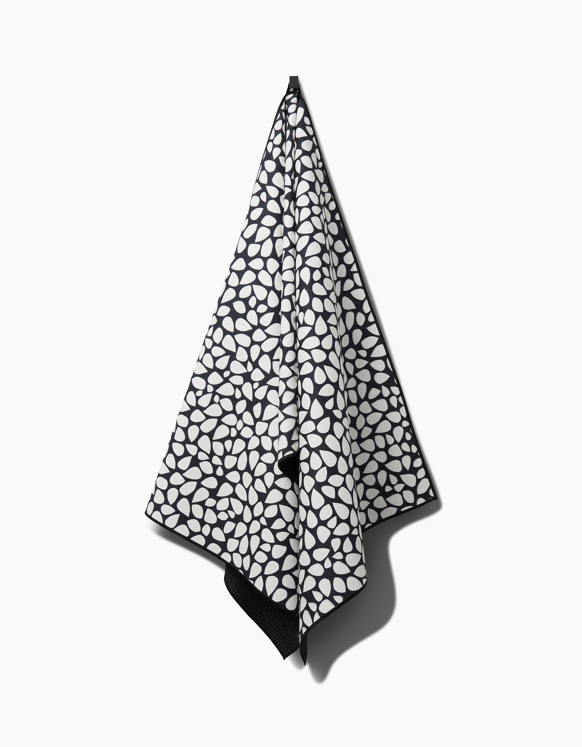 The Pebbles non-slip yoga towel by Marina Solodka features a black and white abstract oval pattern and hangs on a hook against a plain white background.