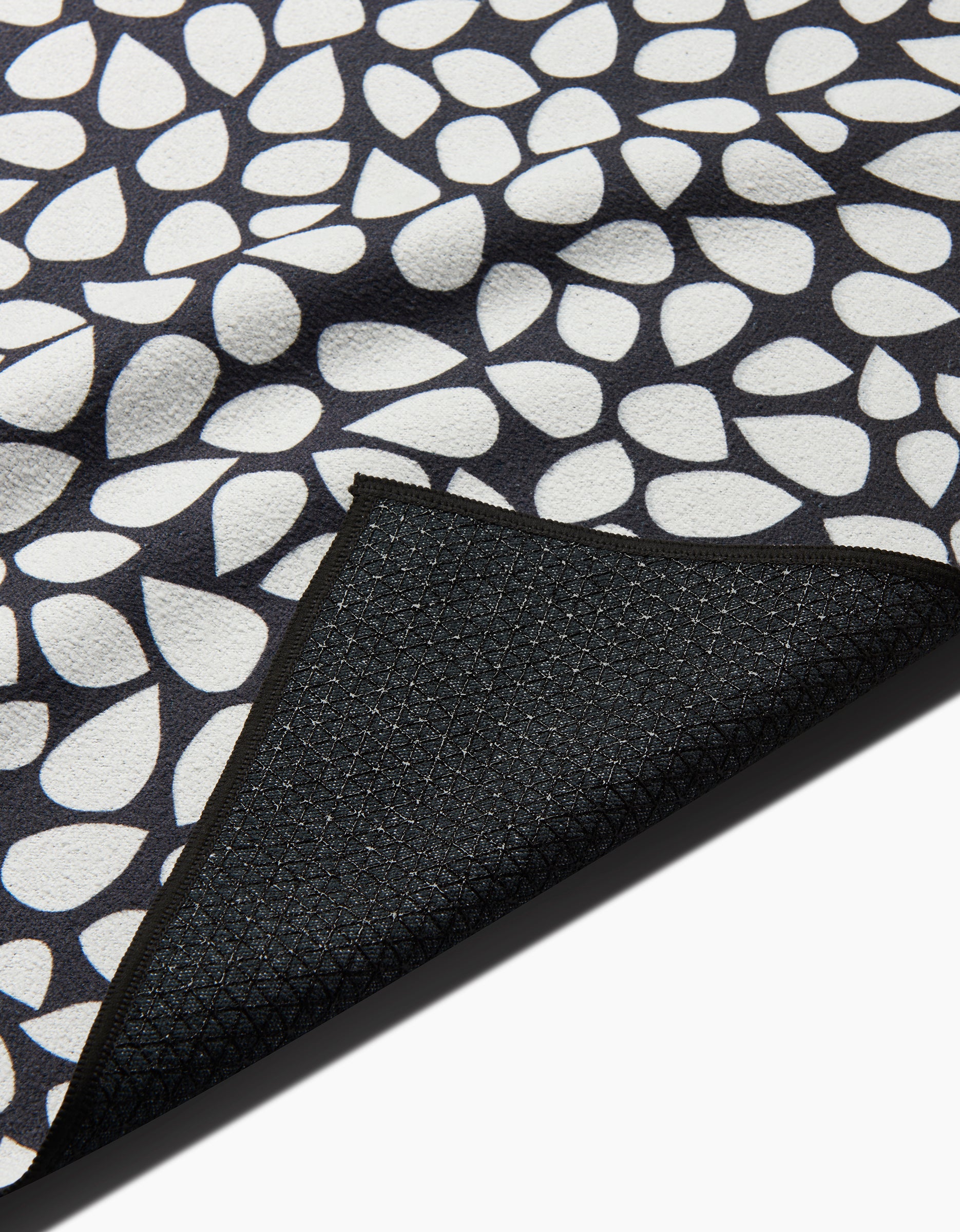 A close-up of the Marina Solodka Pebbles non-slip yoga towel, featuring a white teardrop pattern on black. A folded corner reveals its textured black underside with a subtle grid design.