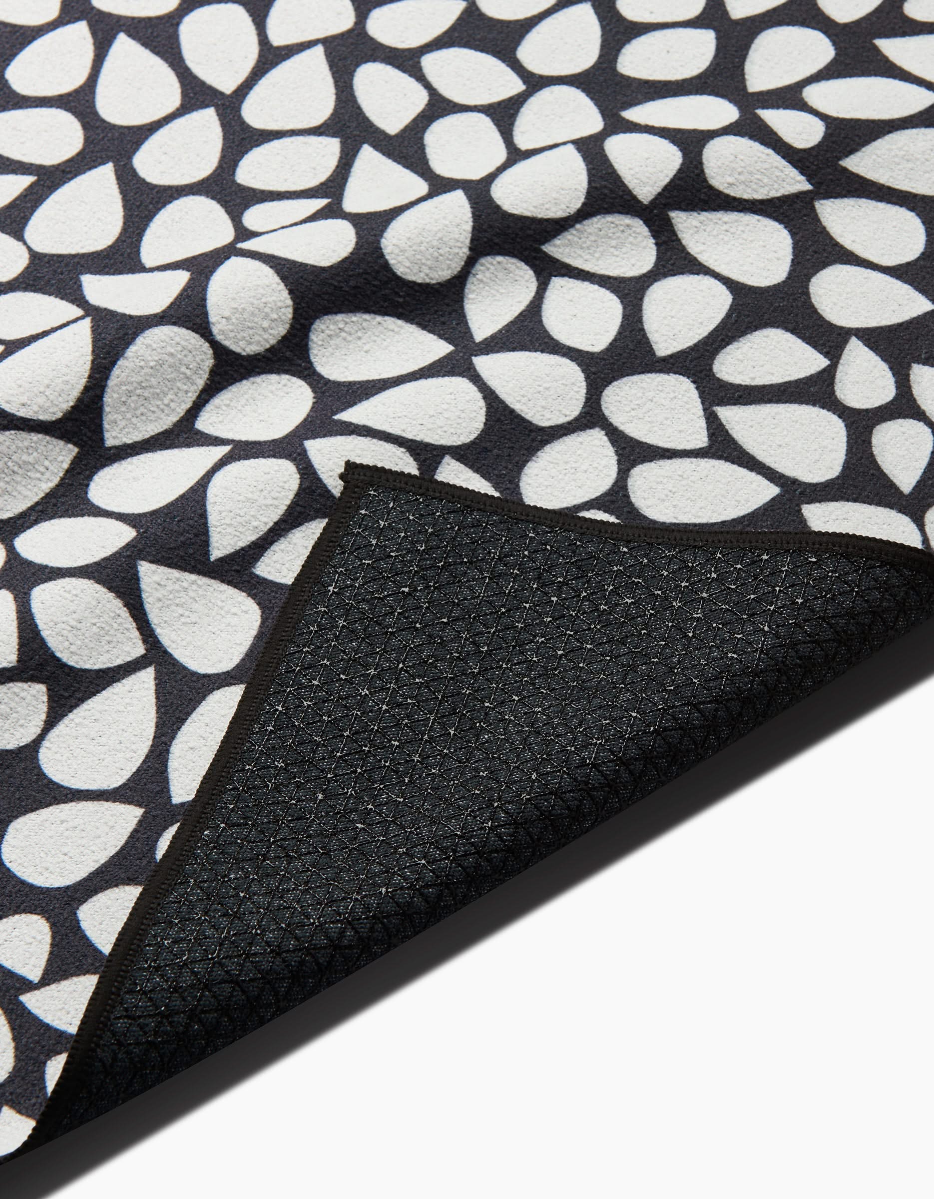 A close-up of the Marina Solodka Pebbles non-slip yoga towel, featuring a white teardrop pattern on black. A folded corner reveals its textured black underside with a subtle grid design.