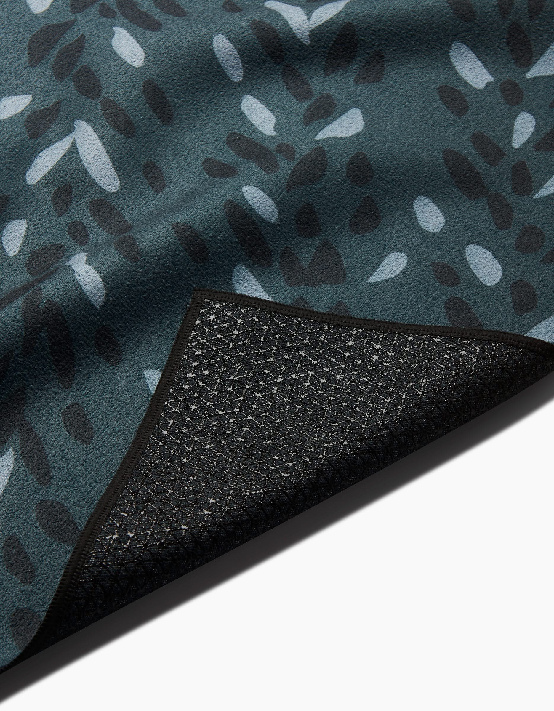 A close-up of the GEOMETRY Pose yoga towel shows its teal textured fabric with a black and white abstract leaf pattern, and a flipped corner revealing the grid-like silicone non-slip grip on the black backing.