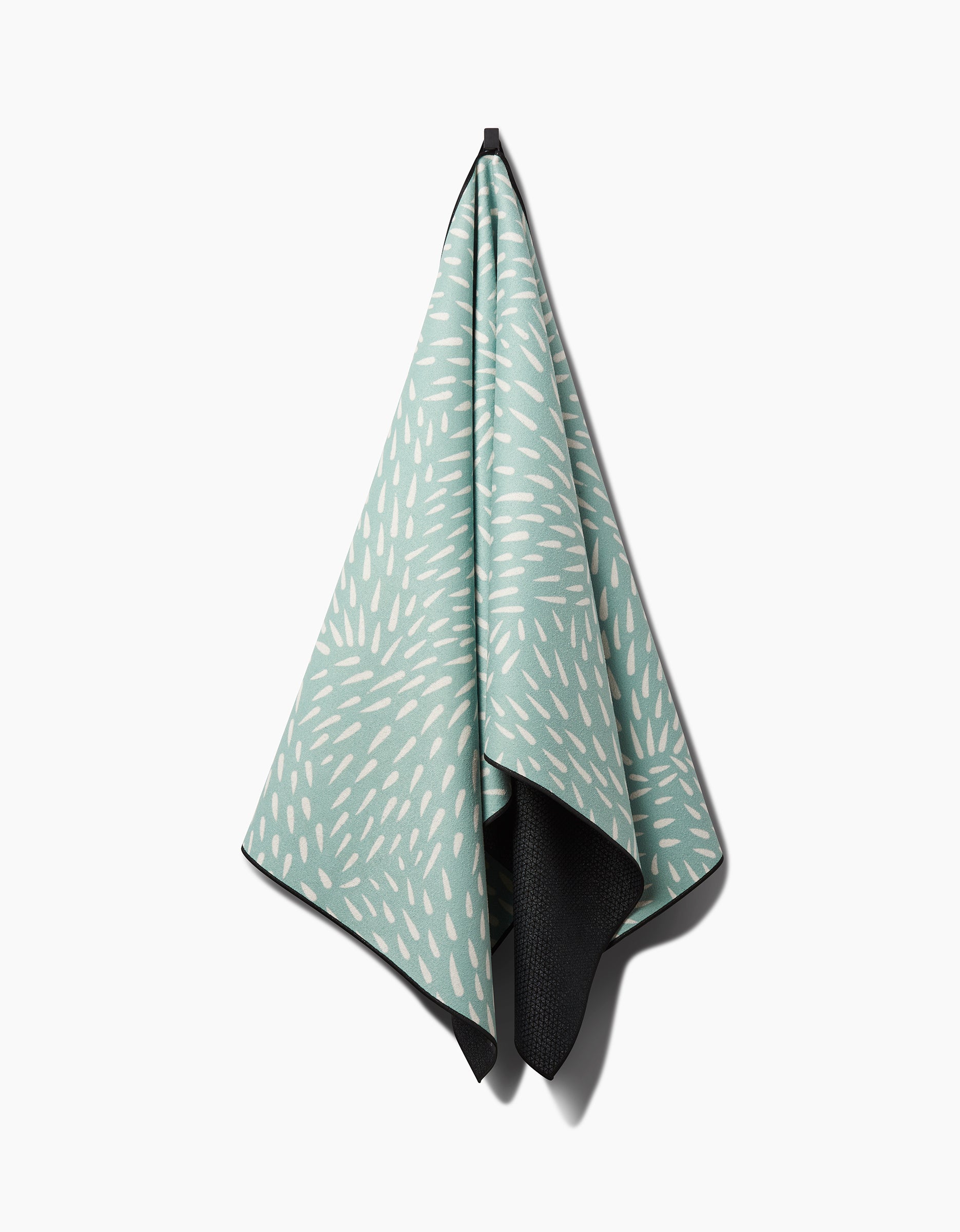 A Rebecca Woolbright Quill Blue yoga towel with white dash patterns hangs from a hook, revealing a corner of its black silicone-grip reverse side, set against a white background.