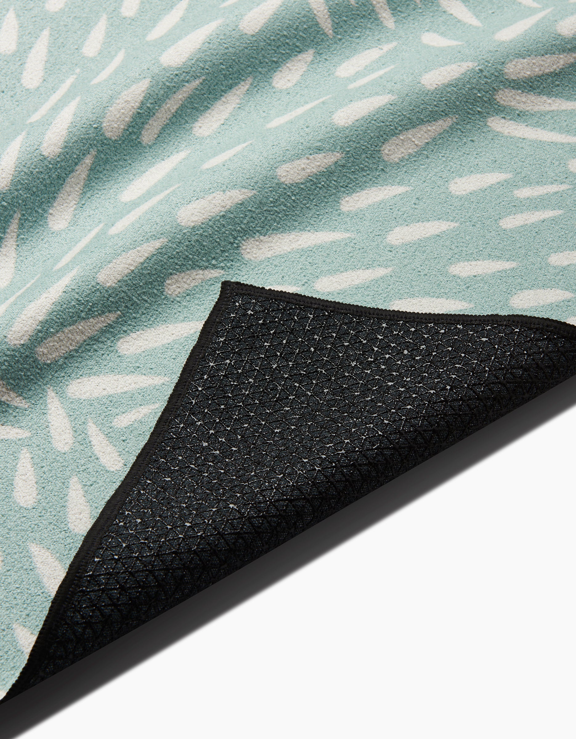 A close-up of the Rebecca Woolbright Quill Blue yoga towel, showing its light blue color with white abstract patterns and a black textured silicone grip non-slip backing folded at one corner.