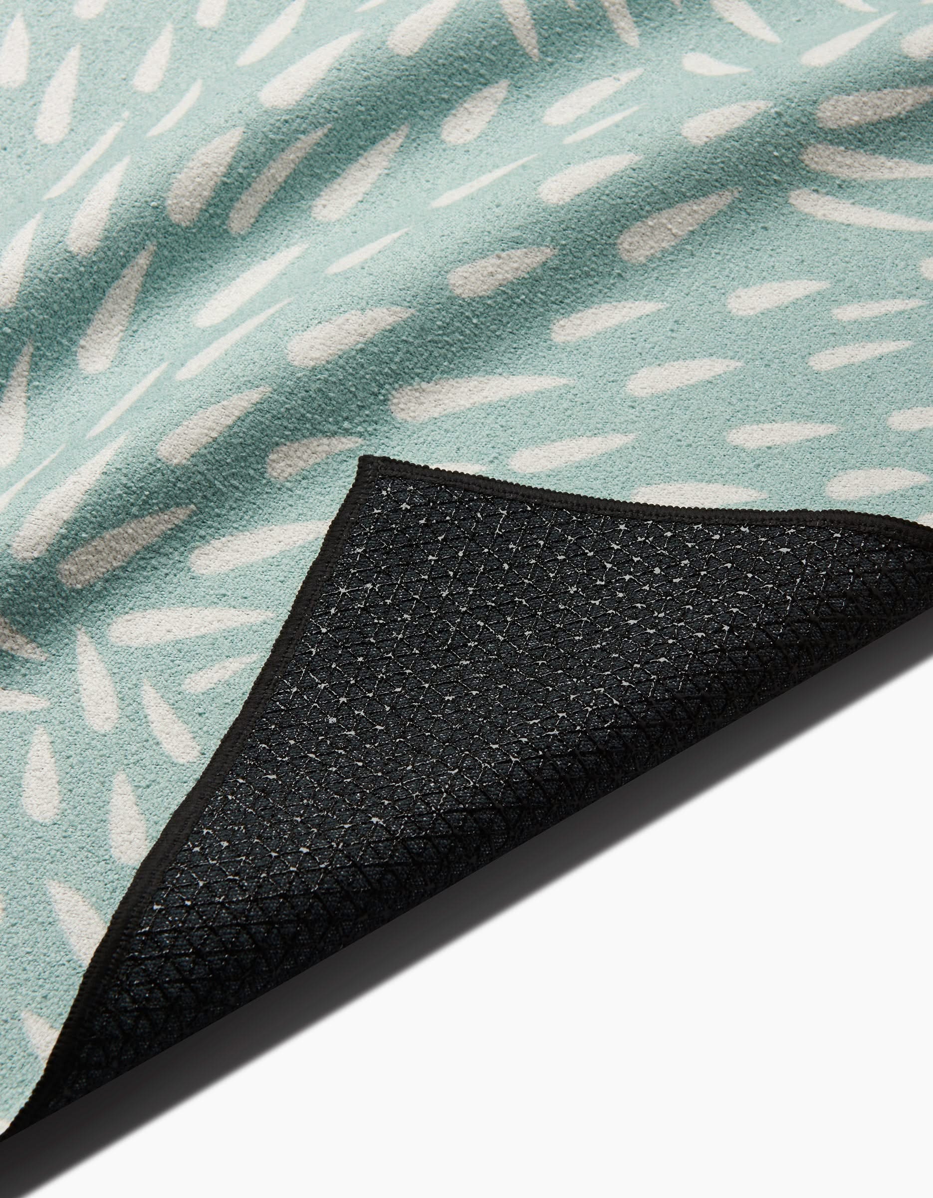 A close-up of the Rebecca Woolbright Quill Blue yoga towel, showing its light blue color with white abstract patterns and a black textured silicone grip non-slip backing folded at one corner.