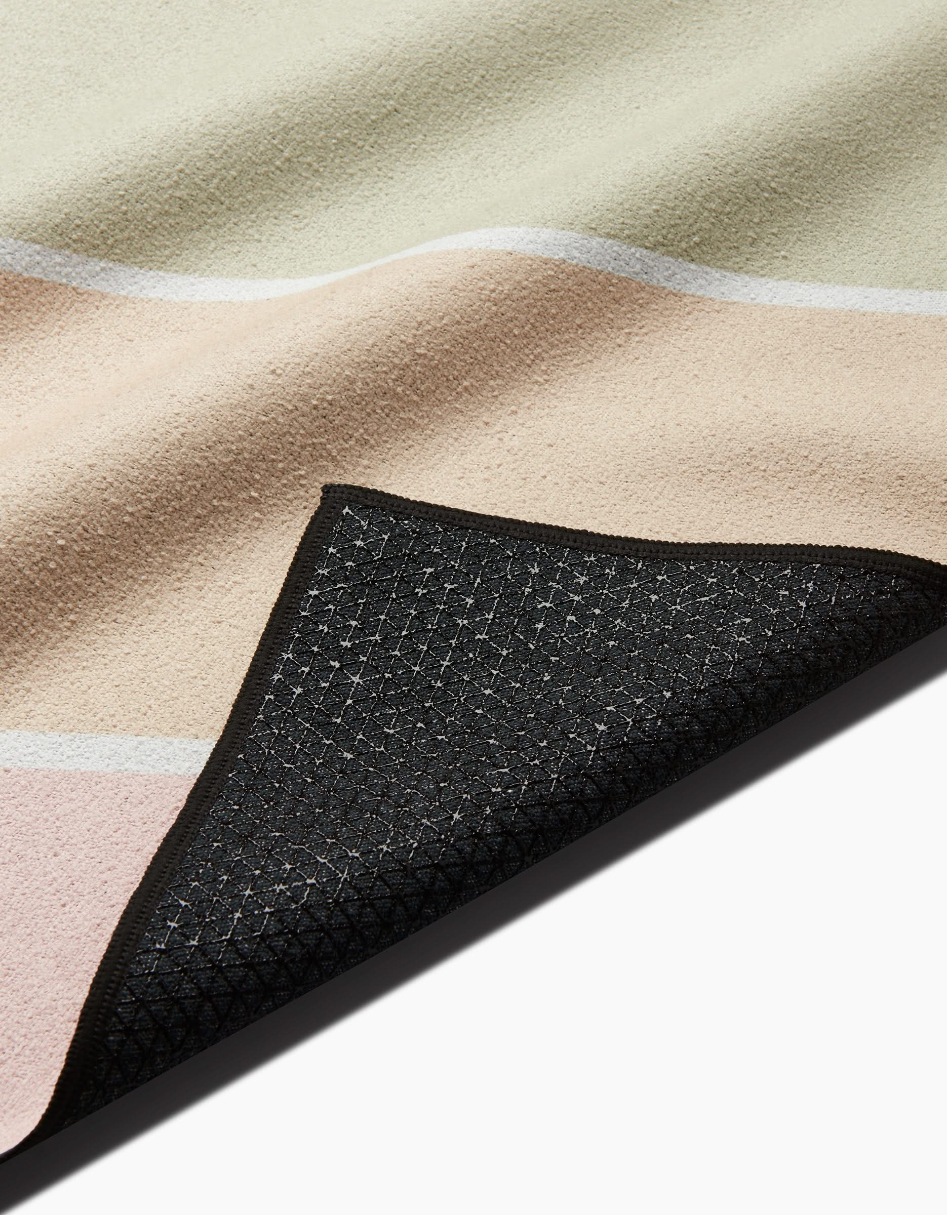 A close-up of the GEOMETRY Rainbow Pastel rug features cream, beige, and blush sections, a textured surface, and black slip-resistant yoga towel-inspired backing with one corner folded over.