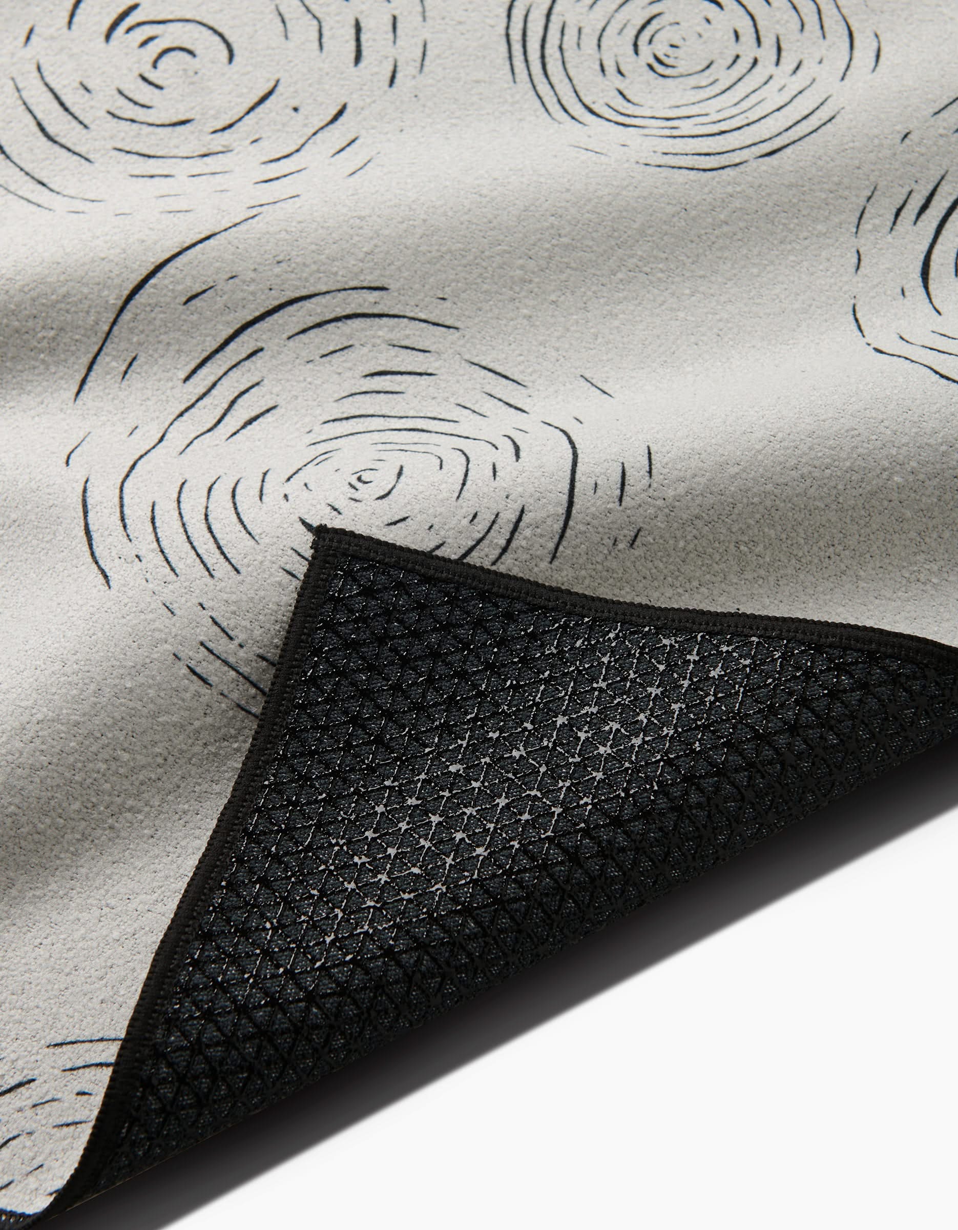 A close-up of GEOMETRY’s Rhythmic yoga towel in light gray with black swirl patterns and a textured black non-slip backing, shown with a corner flipped to reveal the underside.