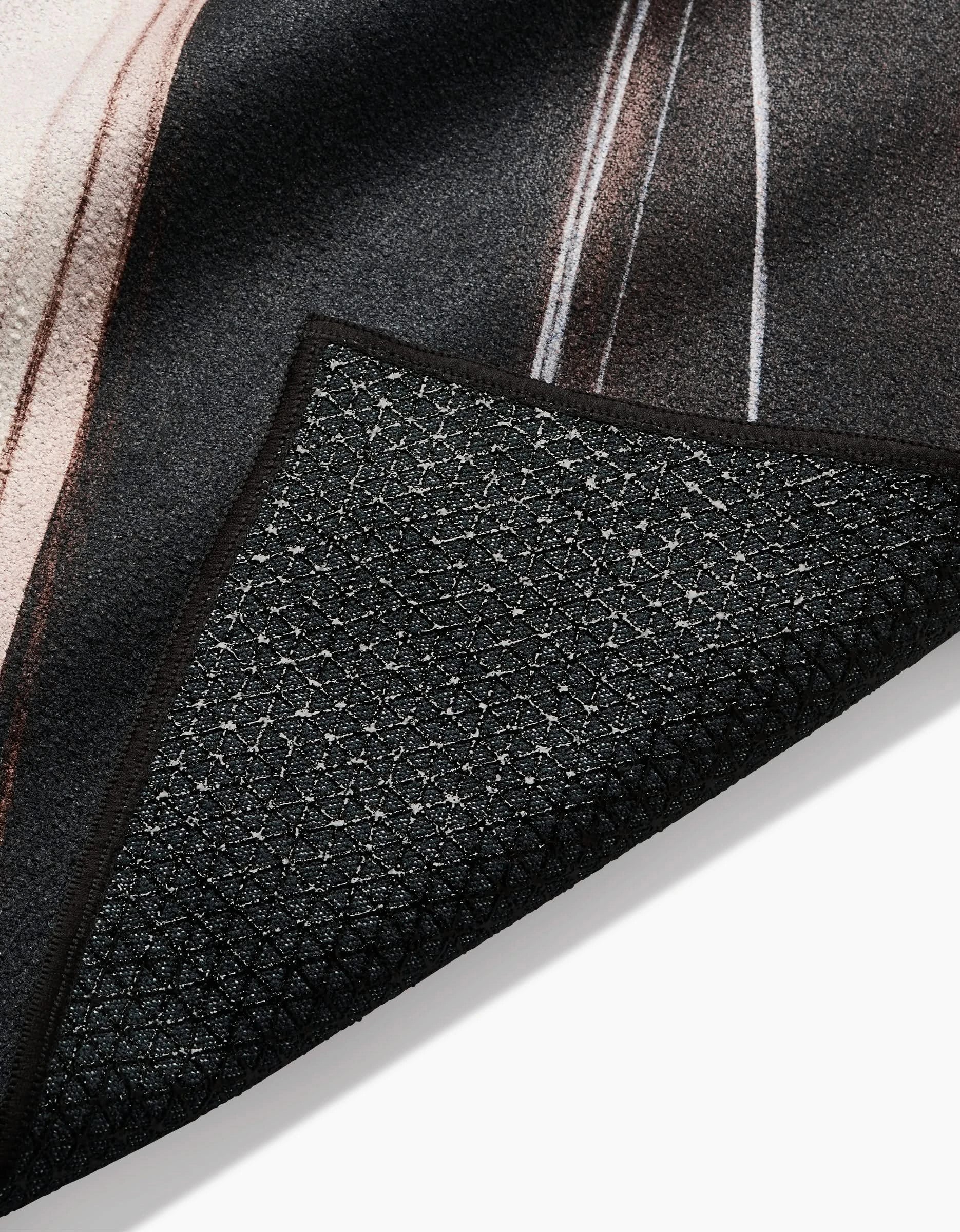 A close-up of a corner of the Seduction yoga towel by Jennifer Janesko, highlighting its black textured non-slip surface, detailed backing underneath, and a glimpse of the patterned top.