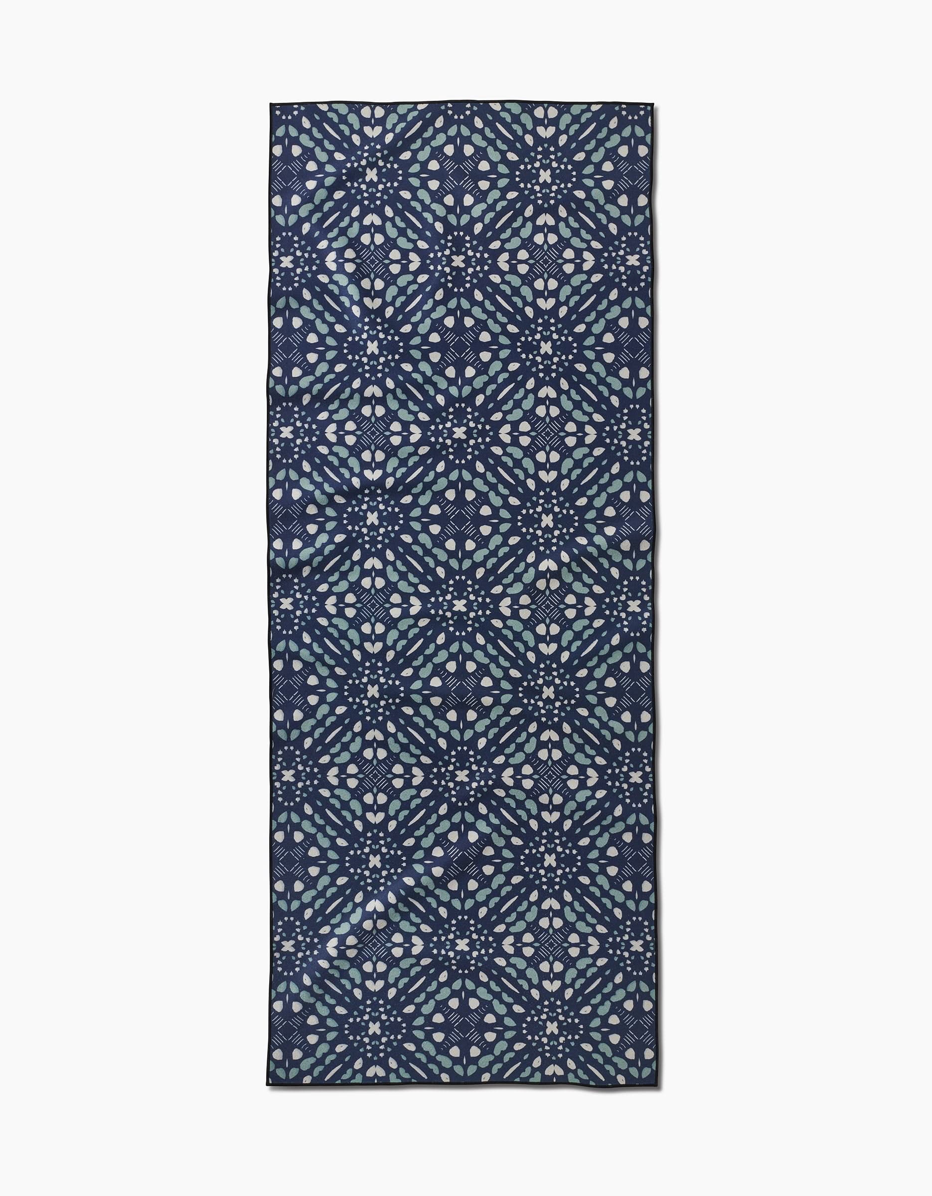 The Twig + Spark Seeds and Nuts yoga towel is rectangular, made from recycled materials, and features a geometric pattern in blue, white, and lavender on a dark background resembling stylized floral star shapes arranged symmetrically.
