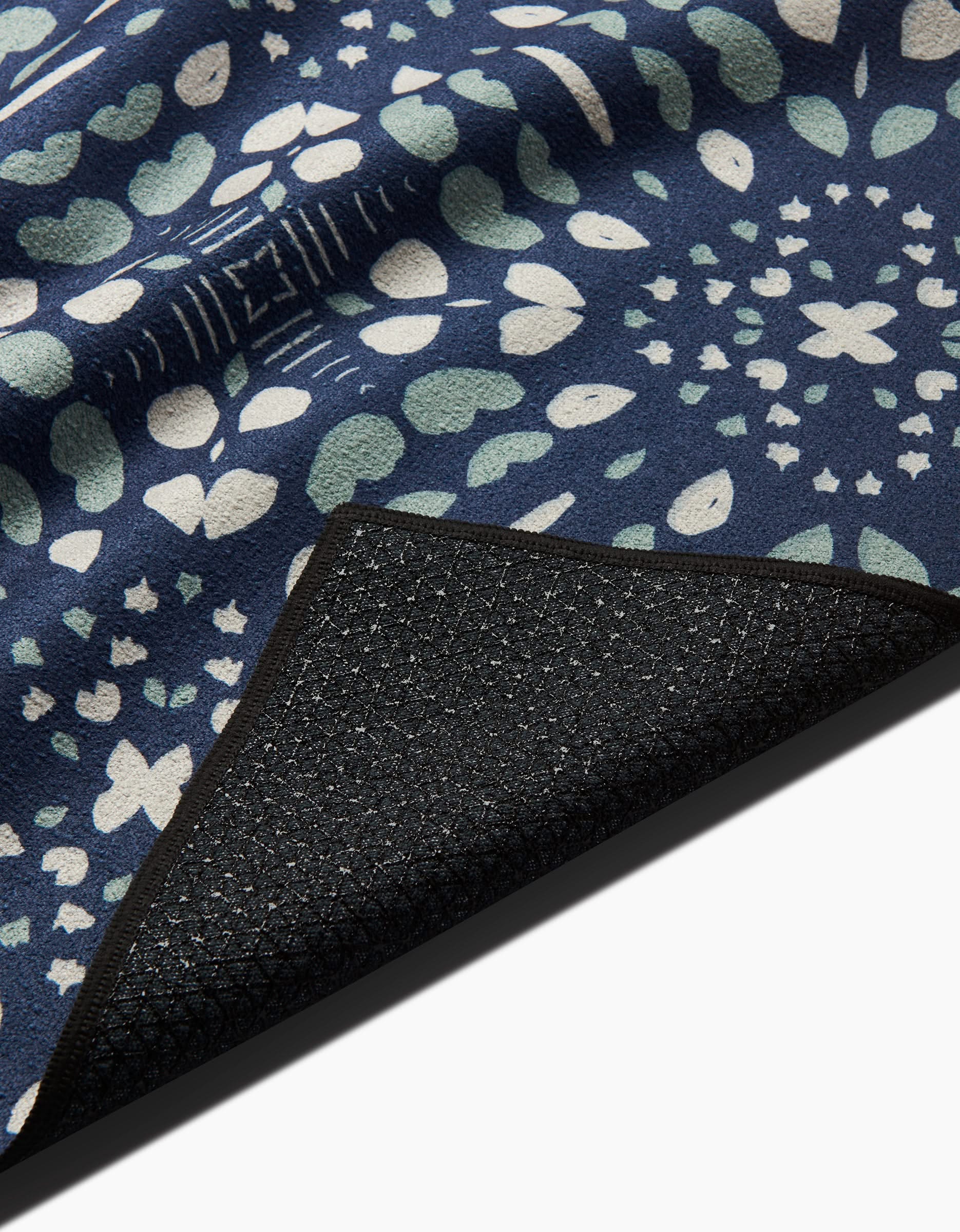 A close-up of the Twig + Spark Seeds and Nuts recycled yoga towel, featuring a blue surface with a white and light green abstract floral pattern; one corner is flipped to show its black textured non-slip backing.