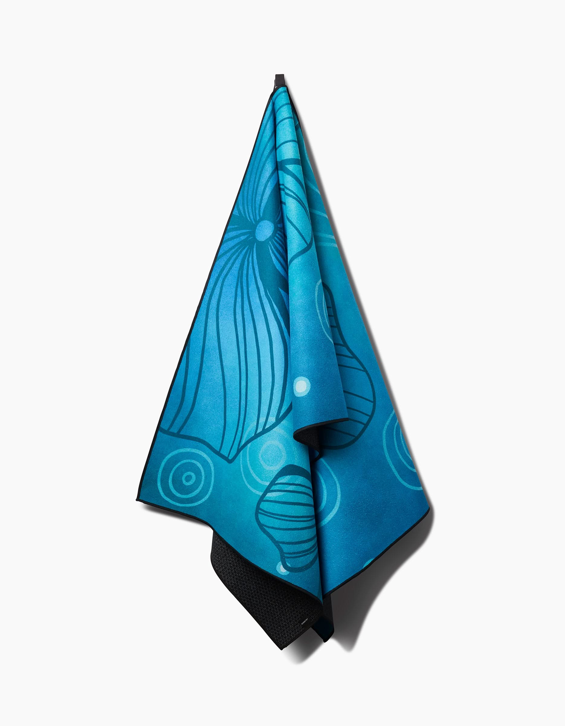 The Blissful Flamingo Serenity yoga towel, in blue with slip-resistant texture, features ocean-inspired abstract patterns of curved lines and circles in lighter blue and white. It’s displayed against a plain white background.