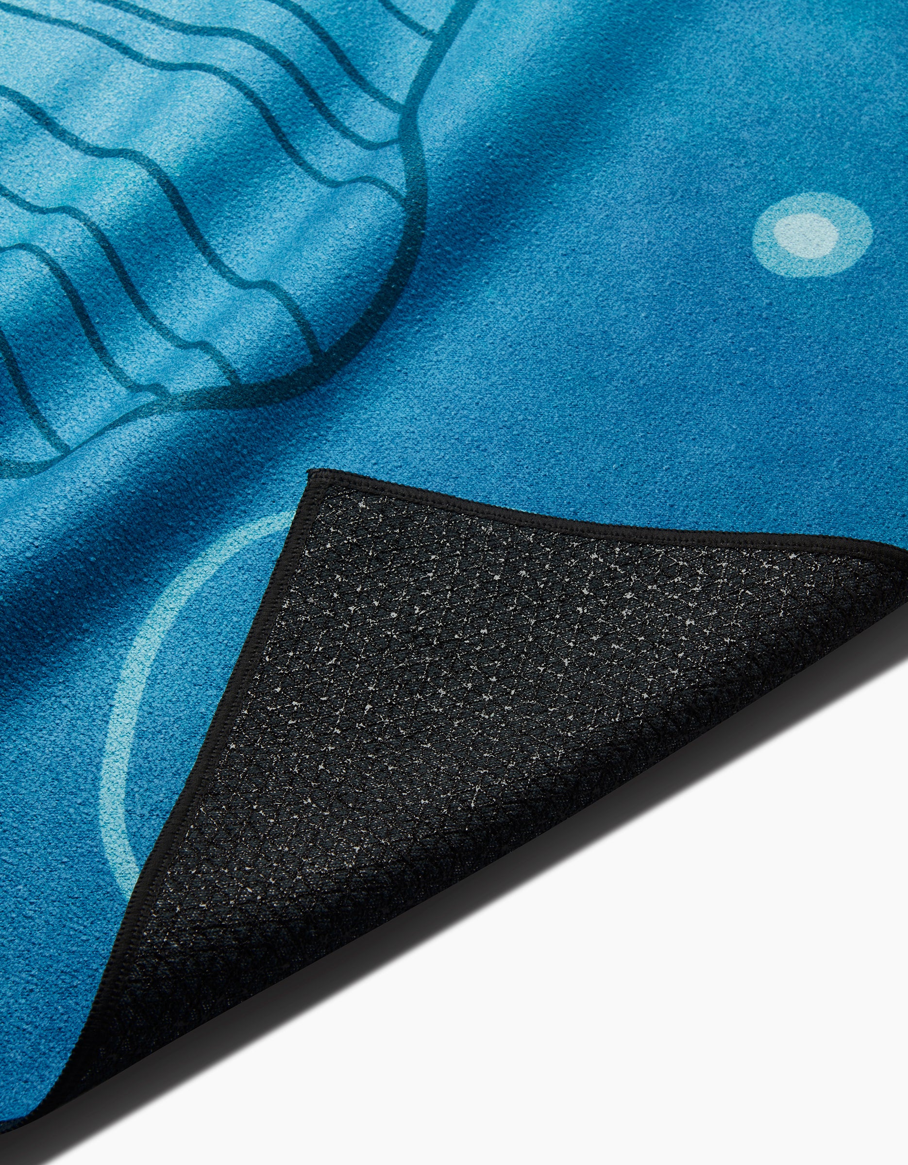 A close-up of the Blissful Flamingo Serenity yoga towel features a wavy blue abstract pattern and a black, textured slip-resistant underside, with the corner flipped up to display both sides.
