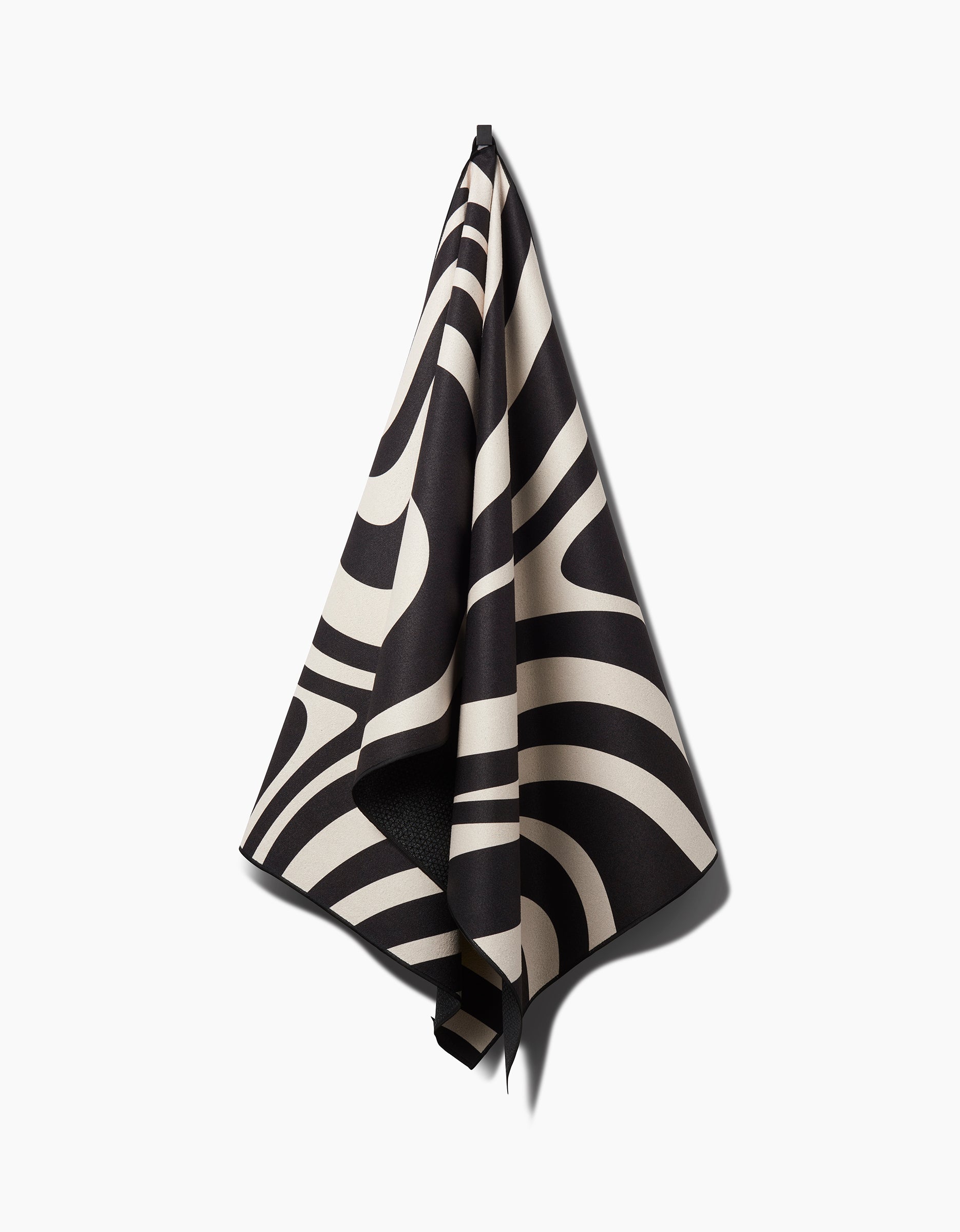 The GEOMETRY Spiral yoga towel features bold black and white abstract curved stripes, hanging on a white wall with a subtle shadow—designed for both style and slip-resistant function during your practice.